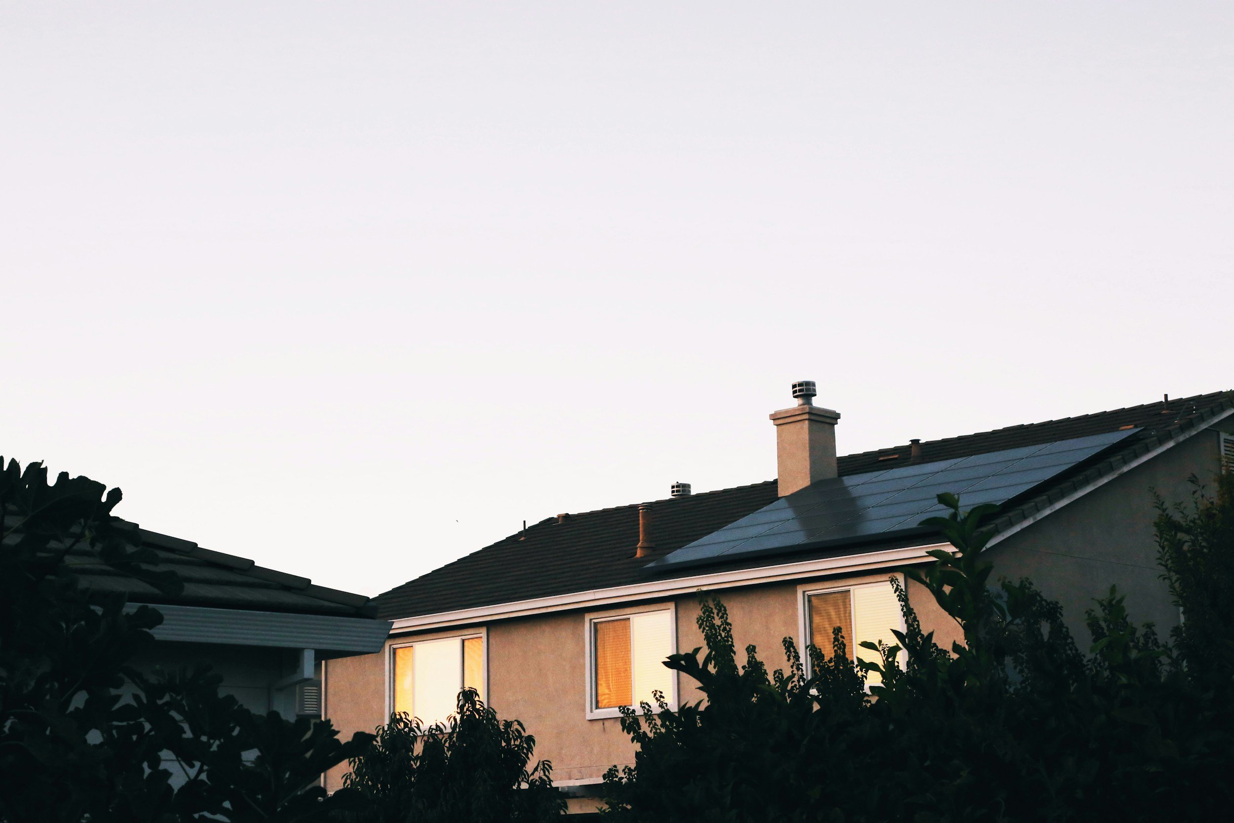 A suburban house with solar panels on the roof, two chimneys, and illuminated windows at sunset, with trees in the foreground.