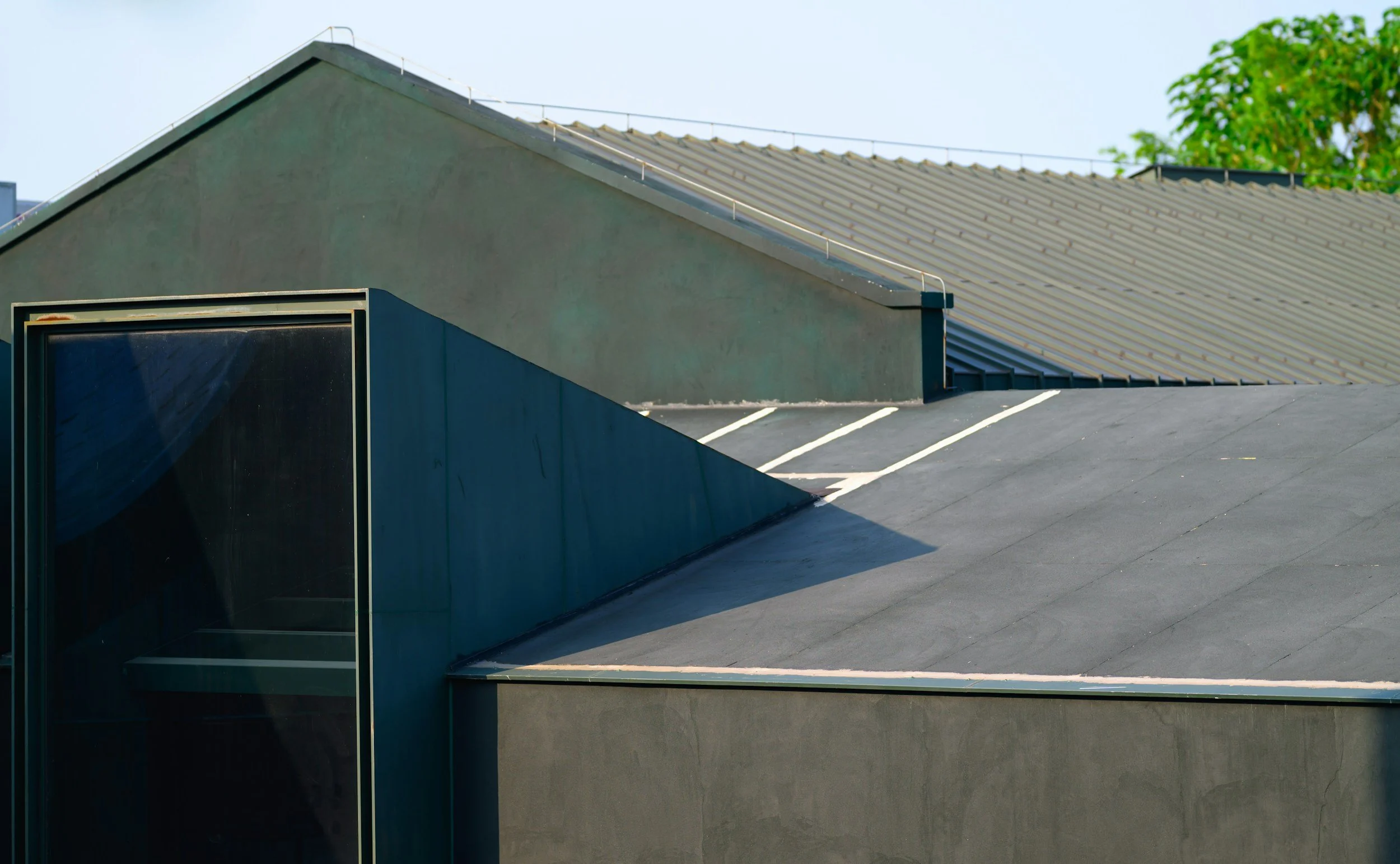 Close-up of a modern building's roof with metal roofing, a small skylight, and a drainage system, clear blue sky, and a green tree in the background.