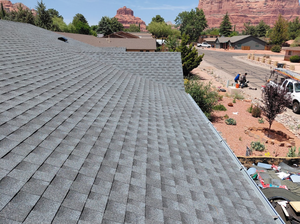 View of a gray asphalt shingle roof with front yard and street below, residential neighborhood with trees and distant red rock formations.