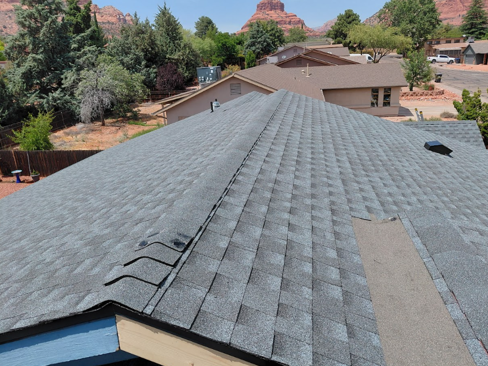 View from a rooftop showing gray asphalt shingles, with some vents and a neighboring house, trees, and distant red rock formations.