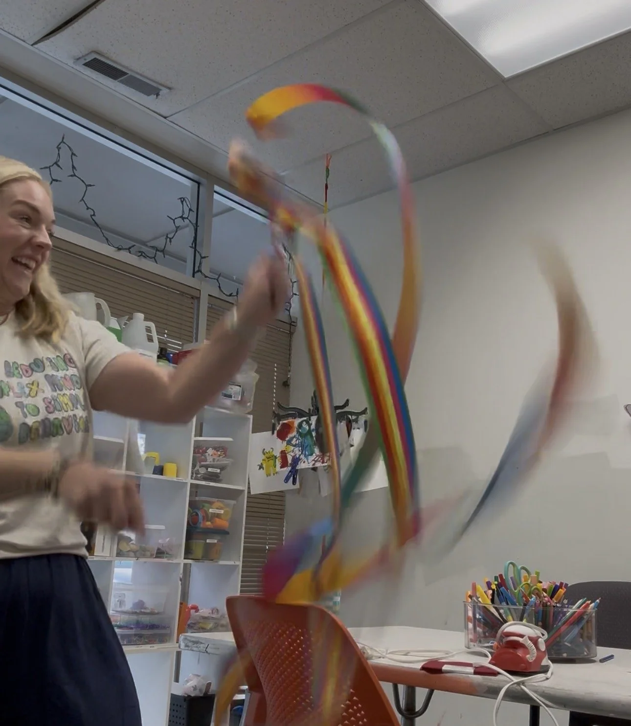 A woman with blonde hair smiling and playing with rainbow-colored streamers in a classroom or craft room setting.