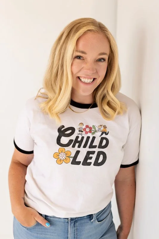 A smiling woman with blonde hair wearing a white T-shirt with black trim. The T-shirt has the phrase 'Child Led' with colorful graphics of children and flowers. She is standing next to a white wall, with one hand in her pocket.
