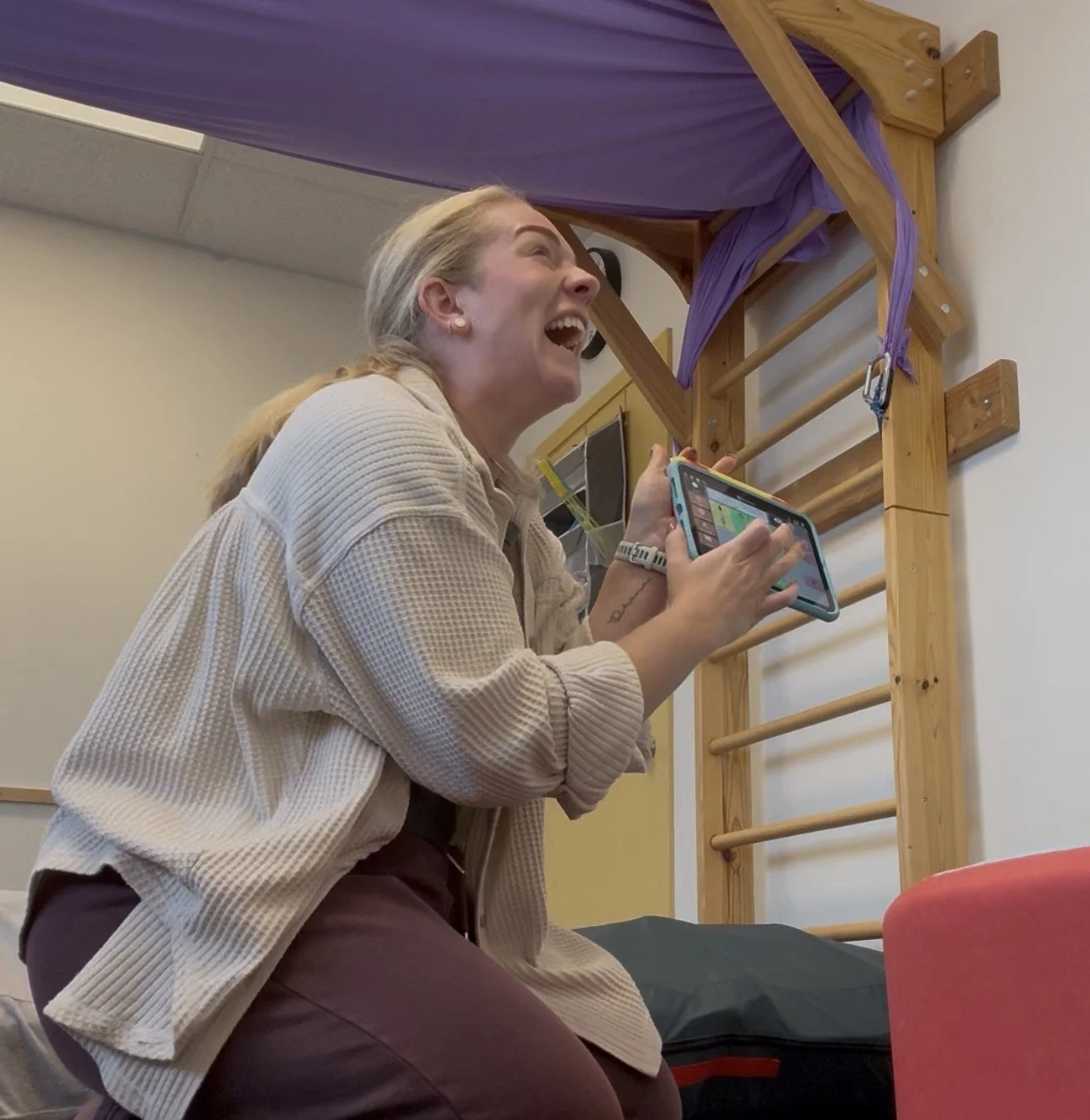 A woman with blonde hair laughing while holding a smartphone, sitting in front of a wooden gym wall ladder with purple exercise bands.