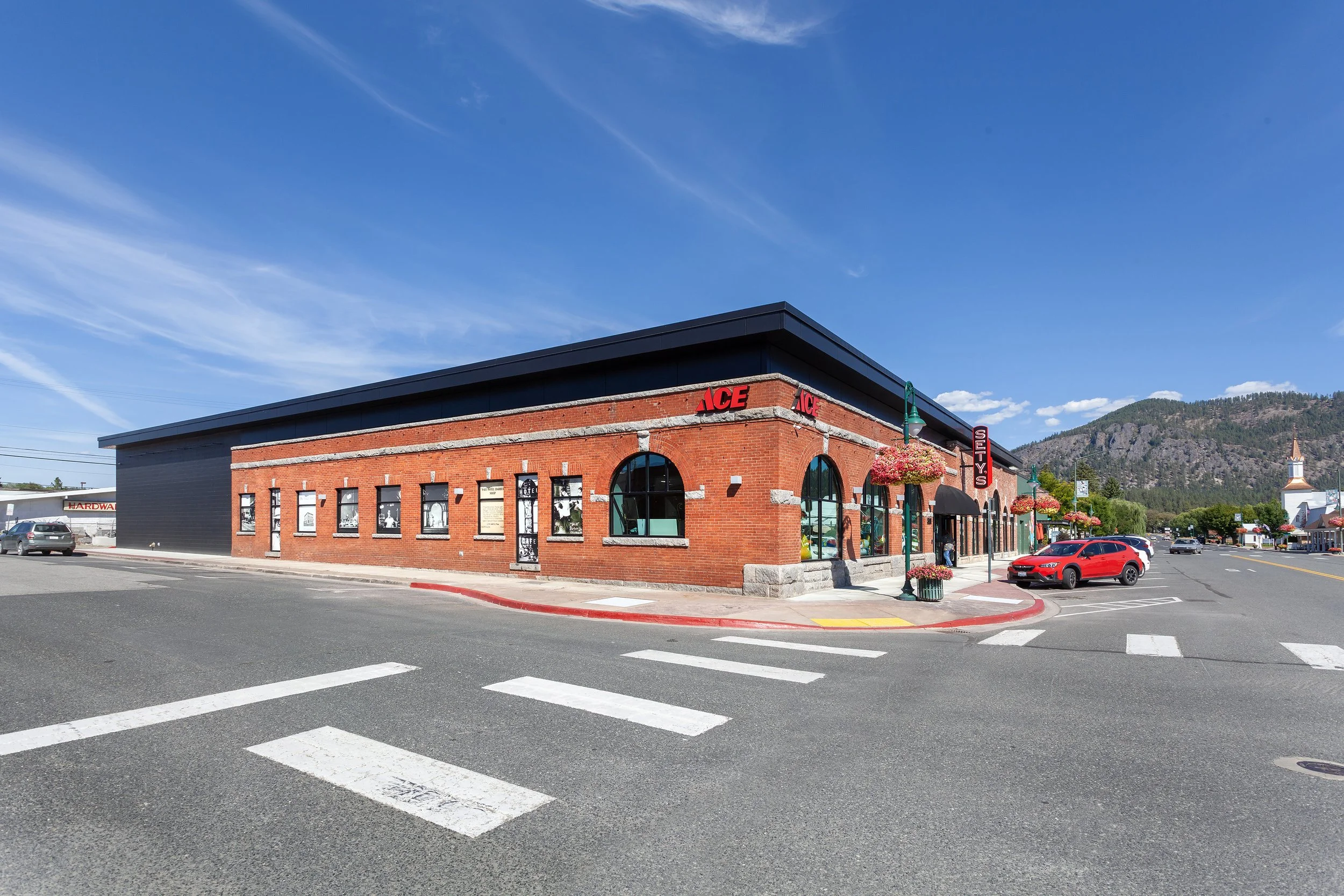 A small brick building with an ACE hardware store sign, parked cars in front, and a mountain in the background under a blue sky with some clouds.