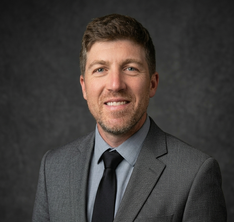 Headshot of a smiling man with light brown hair and beard, wearing a gray suit, light blue shirt, and black tie, against a dark background.
