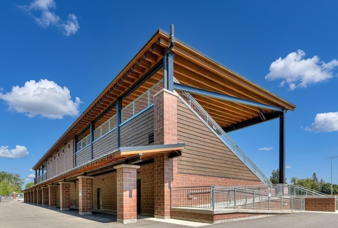 Modern two-story building with brick and wood exterior, large overhanging roof, metal railings, and staircase, under a bright blue sky with clouds.