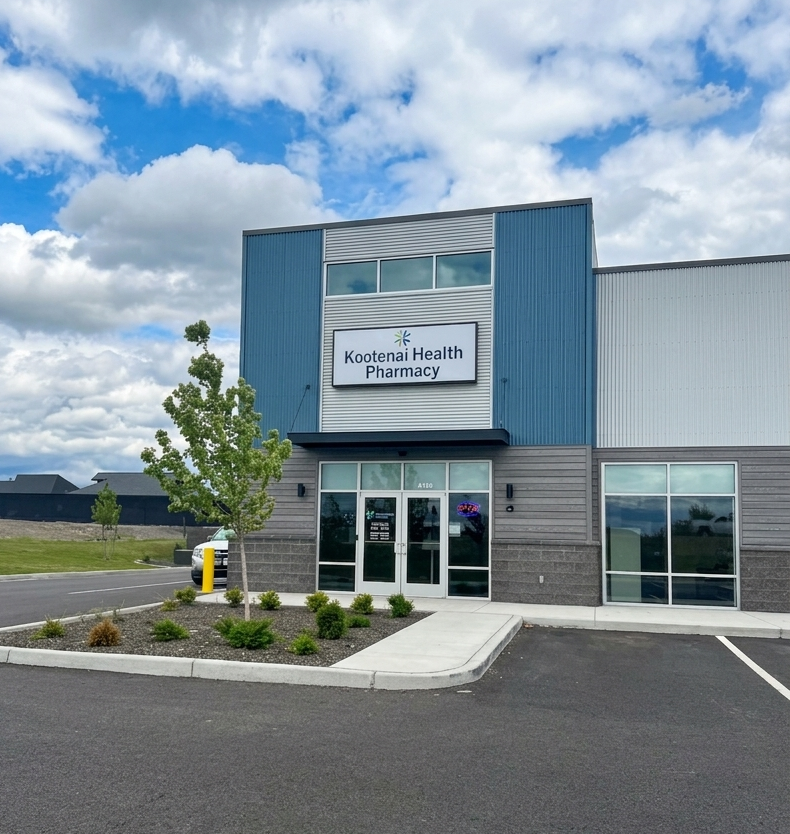 The exterior of Kootenai Health Pharmacy building with a blue and gray facade, glass entrance doors, a small landscaped area with bushes and a tree, and a parking lot in front.