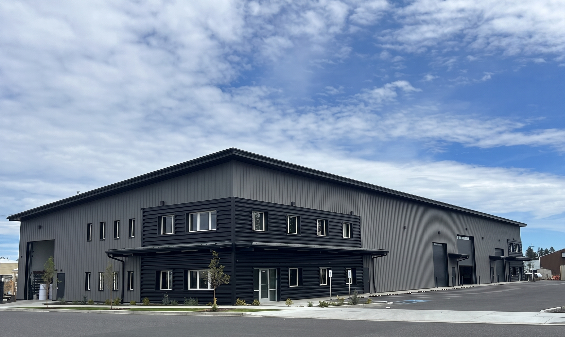 Modern two-story commercial building with gray and black exterior, multiple windows, and a parking lot in front, under a partly cloudy blue sky.