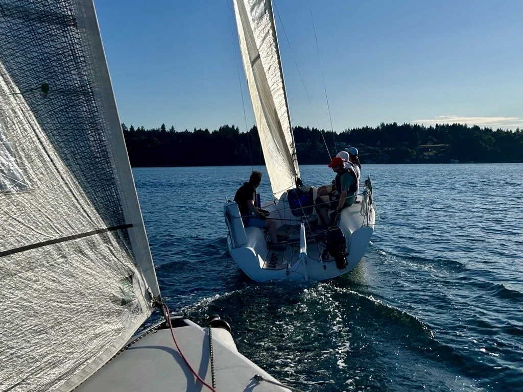A sailboat with people on board sailing on a calm body of water on a sunny day, with a forested shoreline in the background.