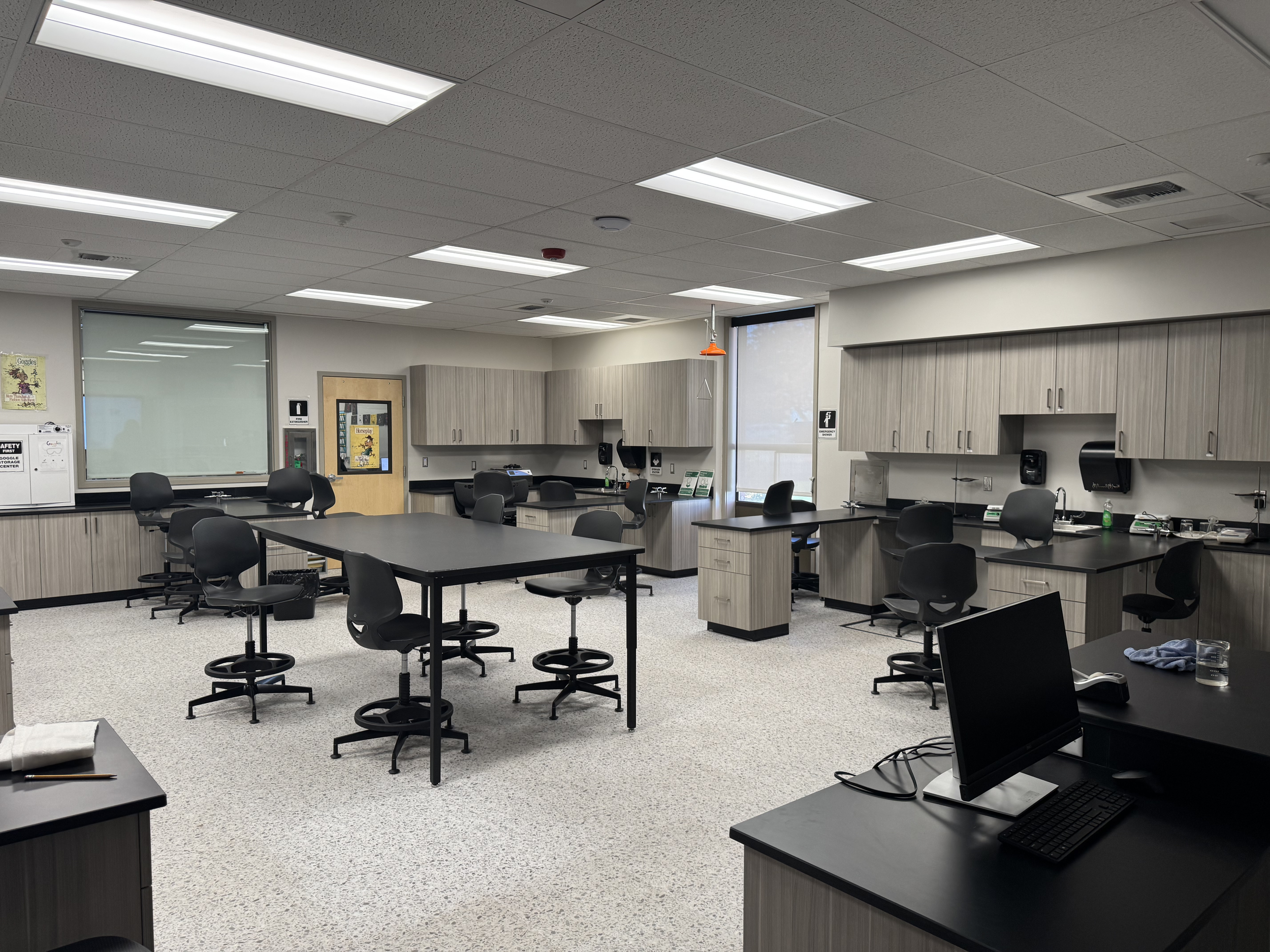 An empty classroom with black desks and chairs, gray cabinets, a whiteboard, and fluorescent ceiling lights.
