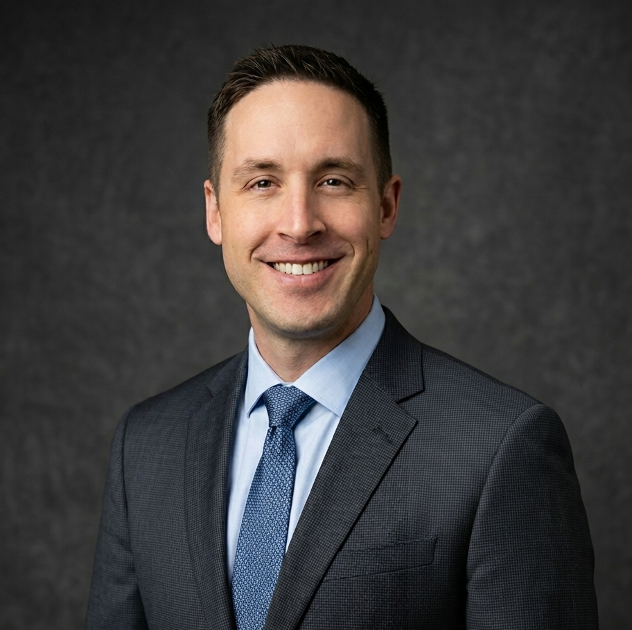 A man wearing a dark suit, light blue shirt, and patterned blue tie, smiling against a gray background.