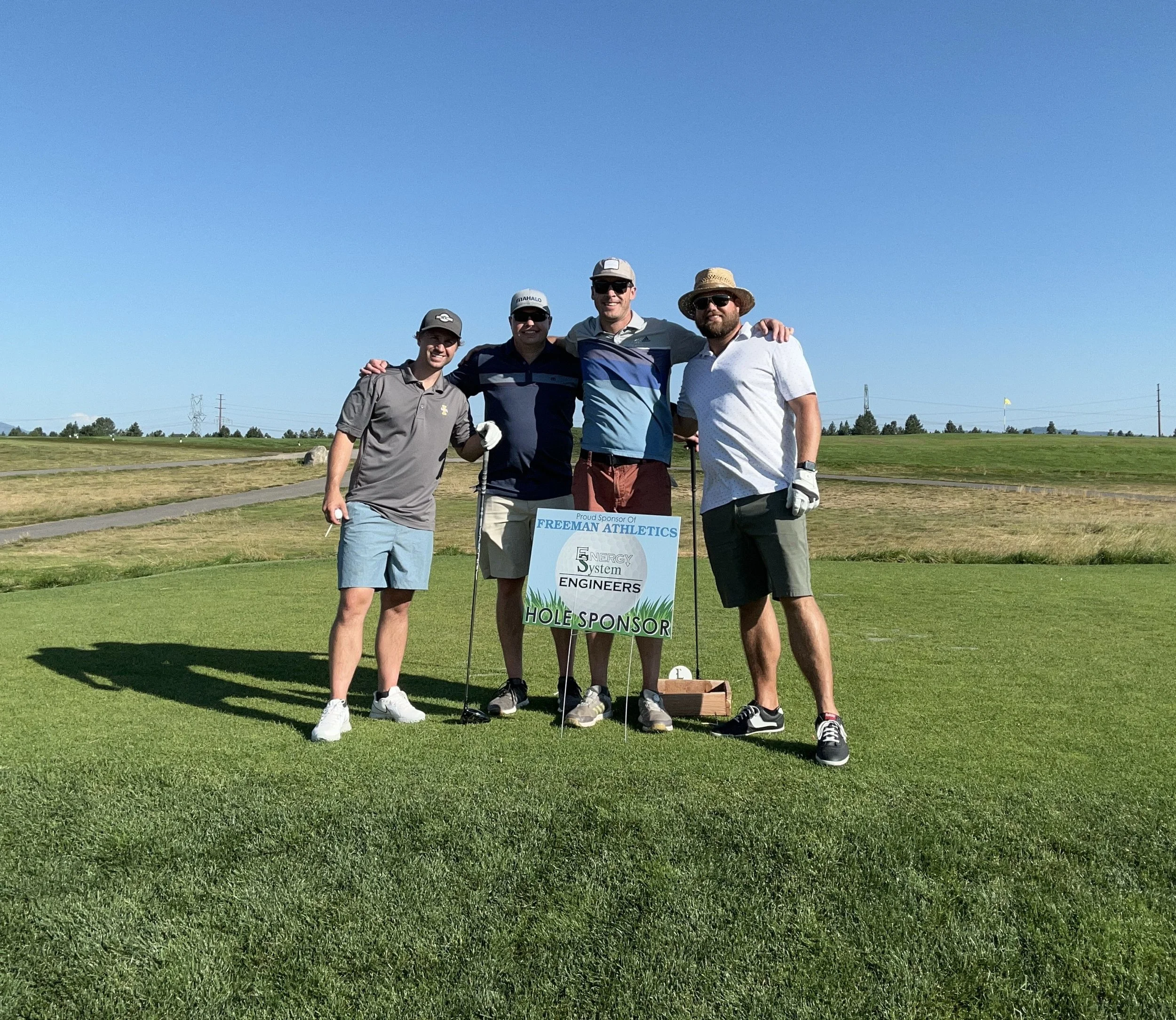 Four men standing on a golf course, holding a sign that says 'Proud Sponsor of Freeman Athletics' with a blue sky background.