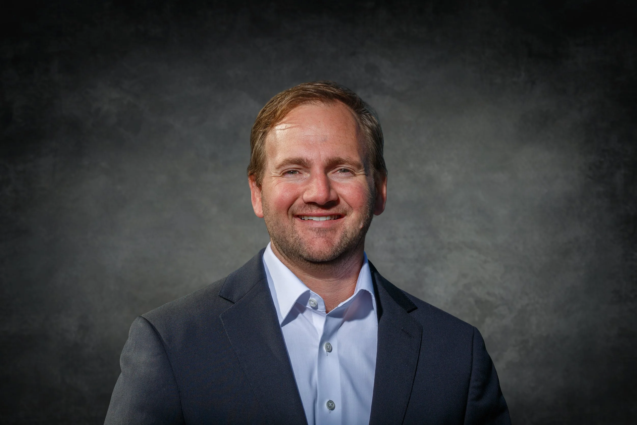 Portrait of a smiling man with light brown hair, wearing a dark suit and white shirt, against a dark textured background.