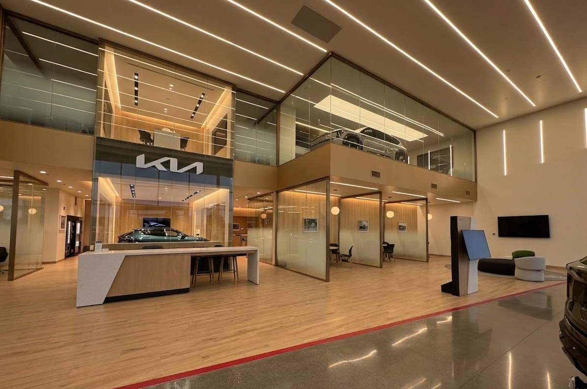 Interior of a car dealership showroom with two stories, showcasing Kia vehicles on the upper and lower levels, lit with modern lighting, with a reception desk, seating area, wall-mounted TV, and financial transaction terminal.