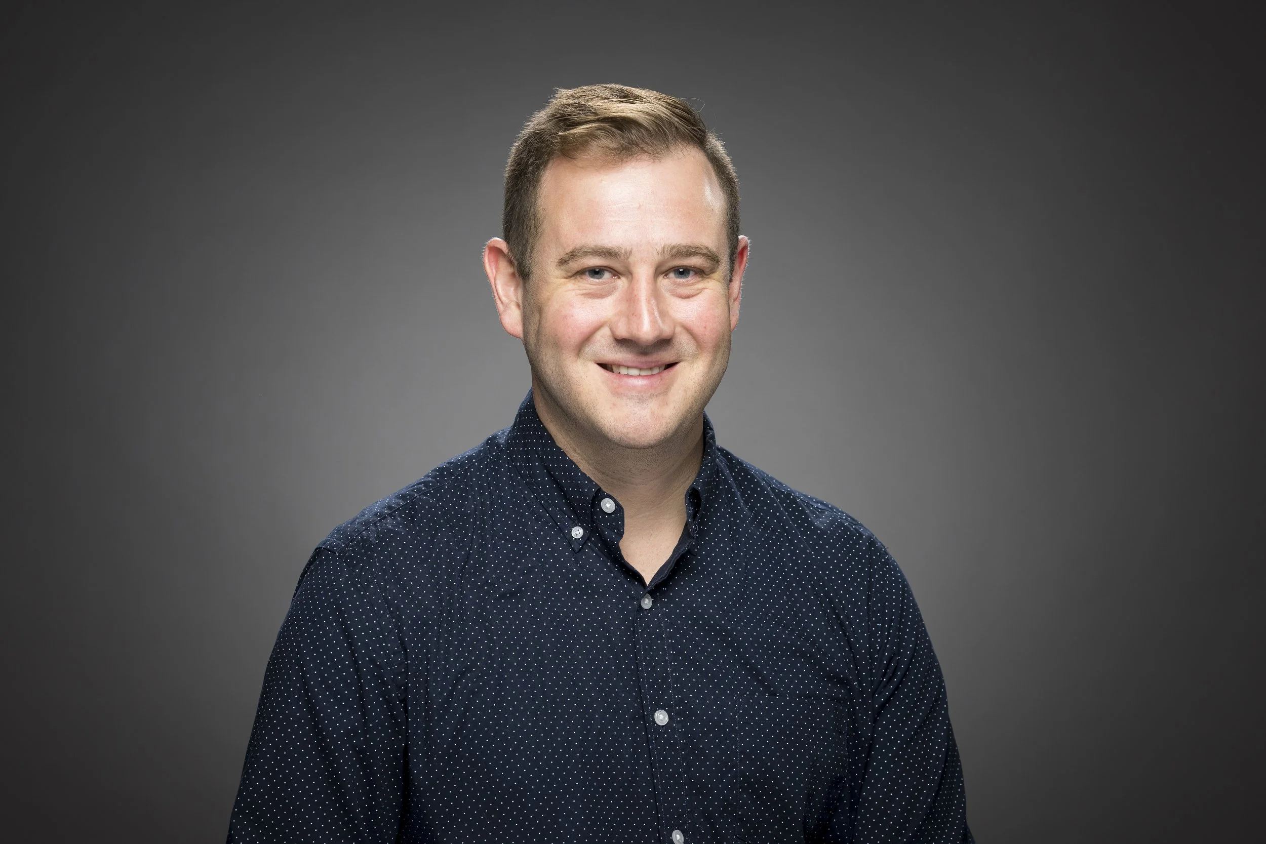 Portrait of a smiling man with short brown hair wearing a dark blue polka-dot shirt against a gray background.