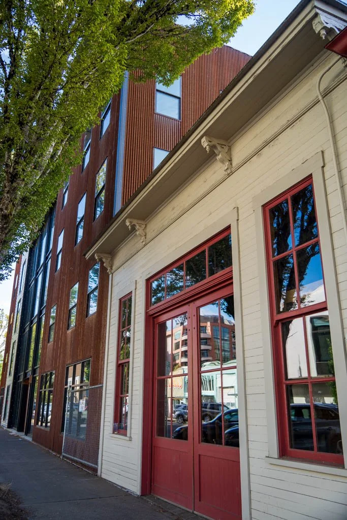 A building with a white exterior and red-framed windows and door next to a modern building with a wooden facade and multiple large windows. A tree with green leaves is partly visible at the top left corner, casting partial shade on the buildings.