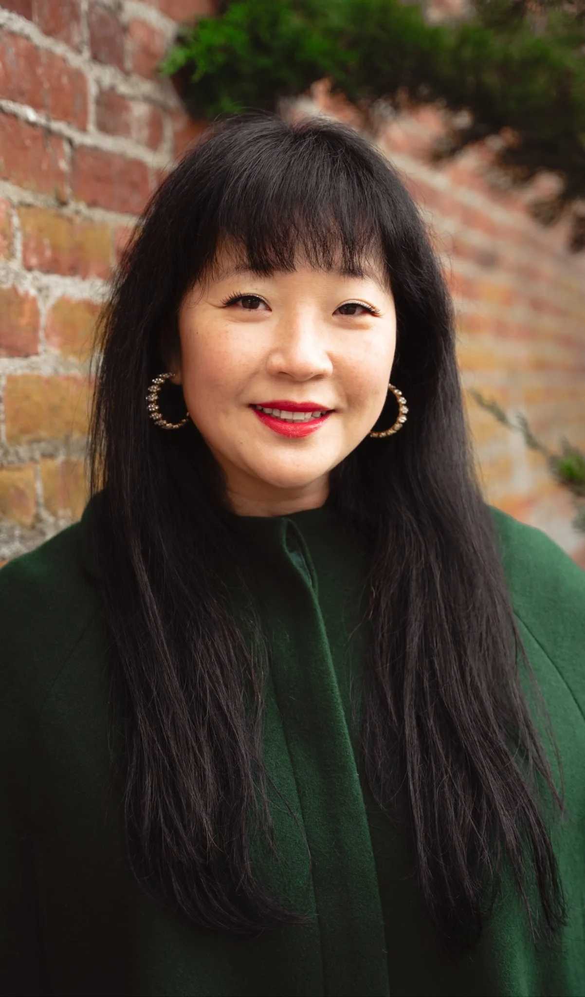 A woman with long black hair, wearing hoop earrings and a dark green top, smiling in front of a brick wall with some greenery.