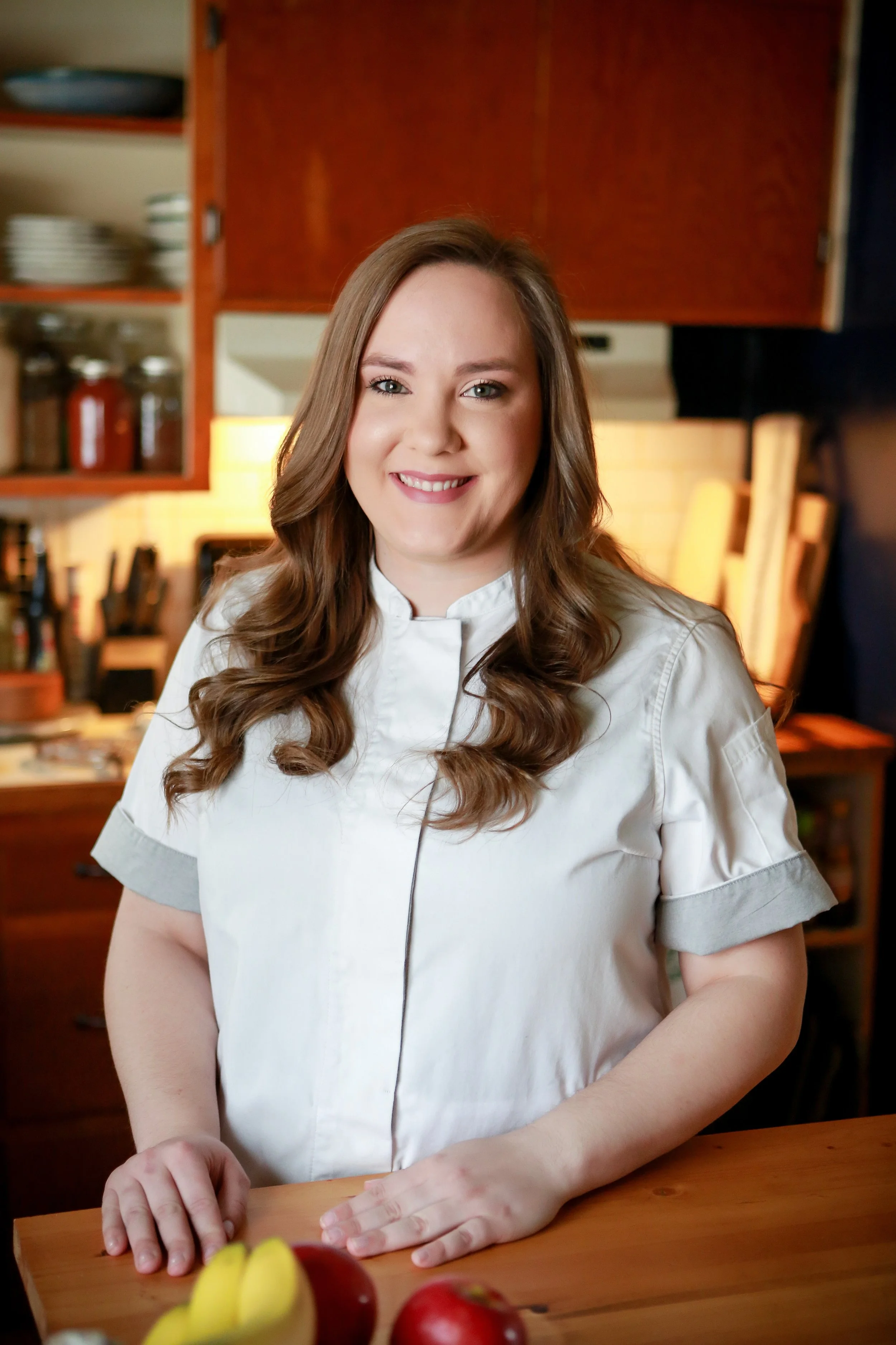 A woman in a white chef's jacket standing in a kitchen, with wooden cabinets and shelves of dishes in the background.