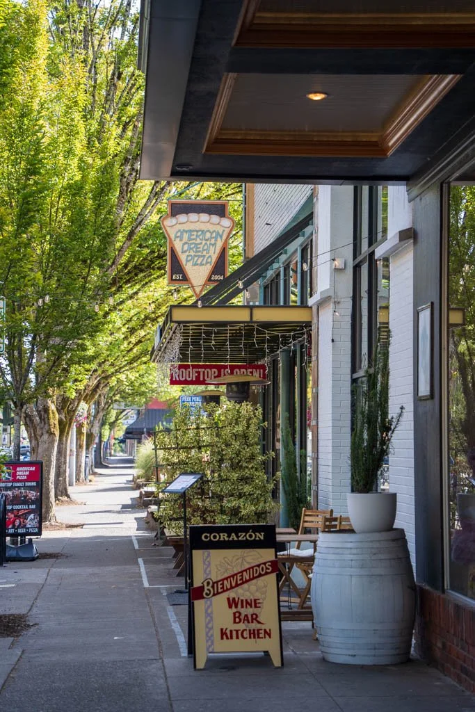 Sidewalk view of a restaurant with signs saying 'American Dream Pizza,' 'ROOF TOP IS OPEN,' and 'CORAZÓN Bienvenidos Wine Bar Kitchen,' with trees lining the street.