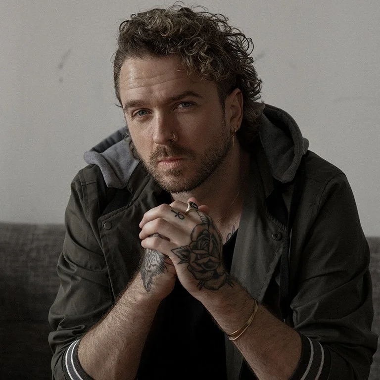 A man with curly hair and a beard, wearing a black jacket with a gray hood, sitting on a couch, with his hands clasped together in front of him.