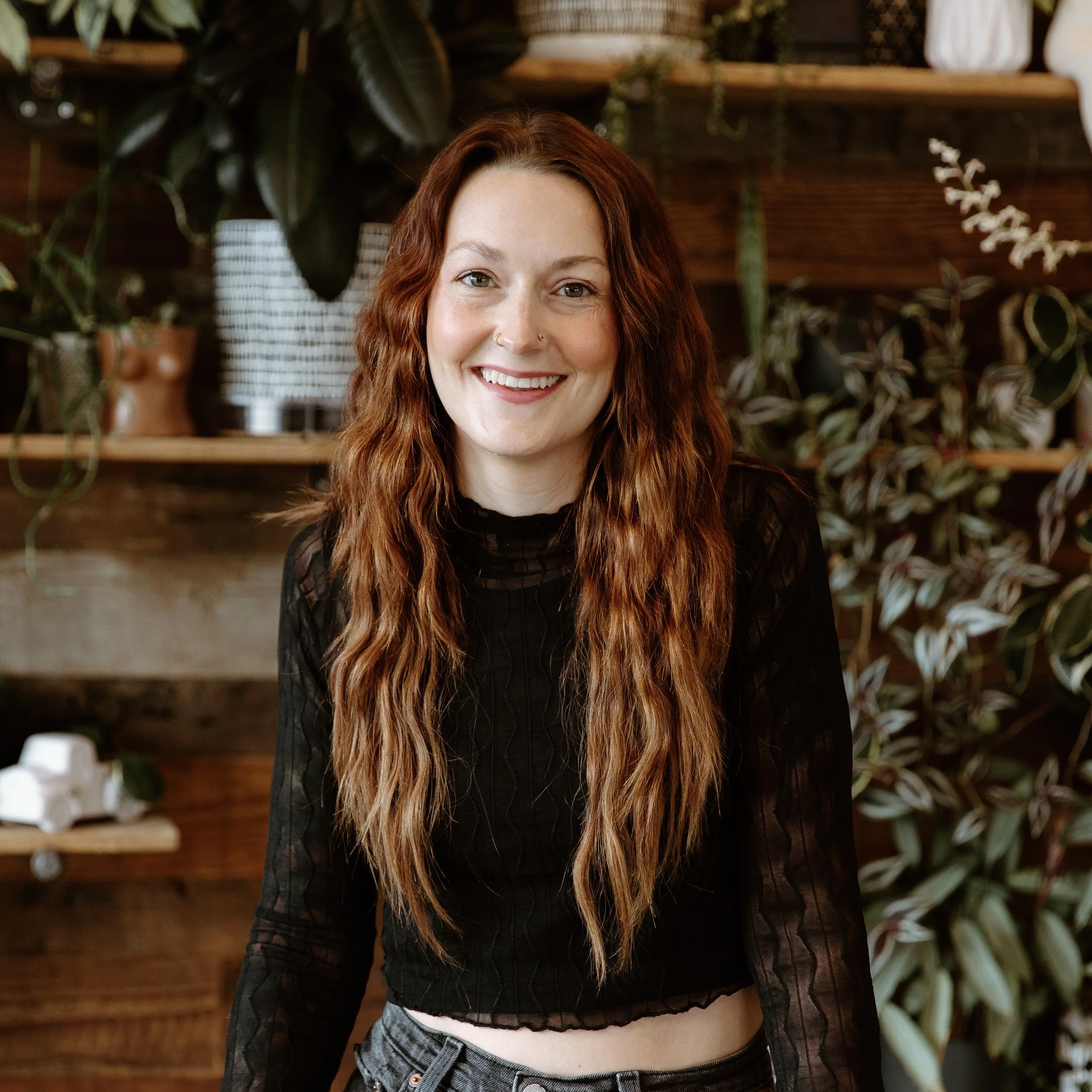 A young woman with long, wavy red hair and light skin smiling indoors in front of a background with plants and wooden shelving.