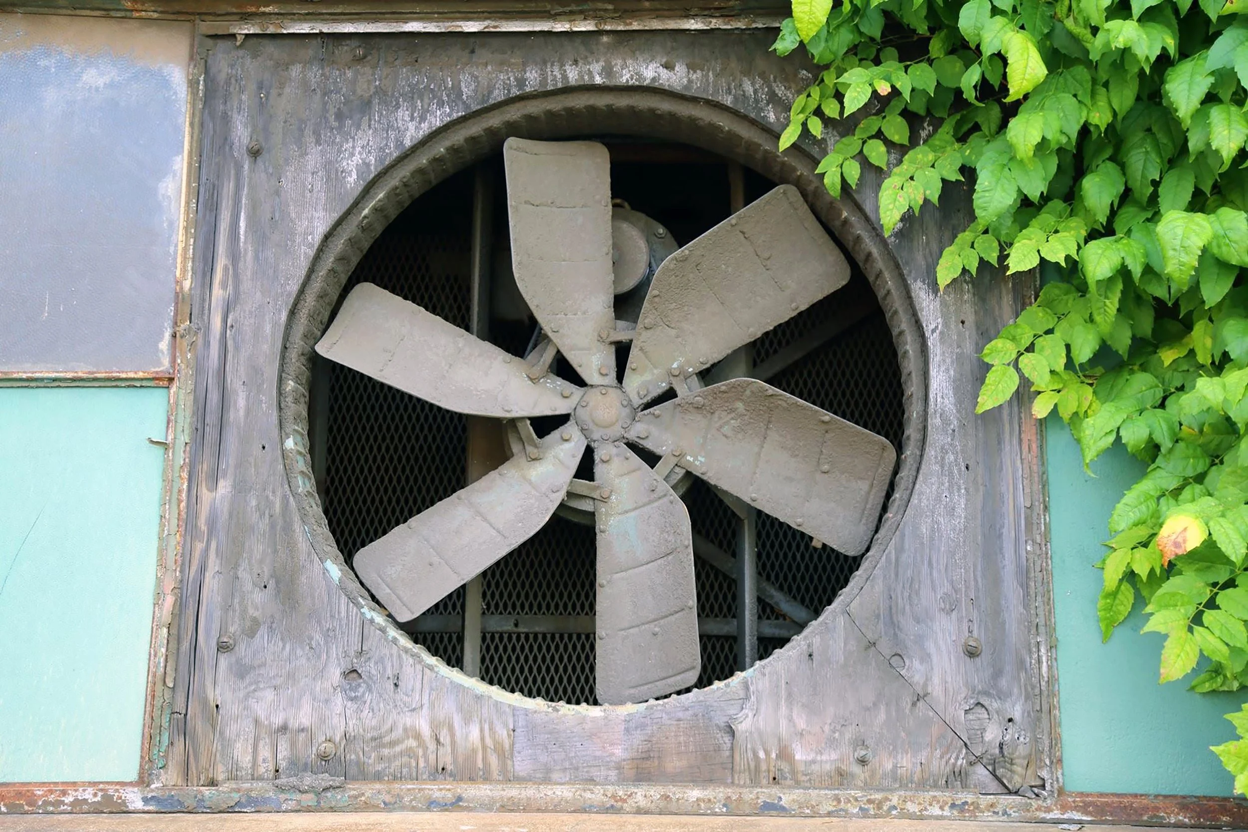 An old, weathered industrial ventilation fan with a large metal blade выполнено не рендером, surrounded by wooden and metal structures, with green foliage growing around it.
