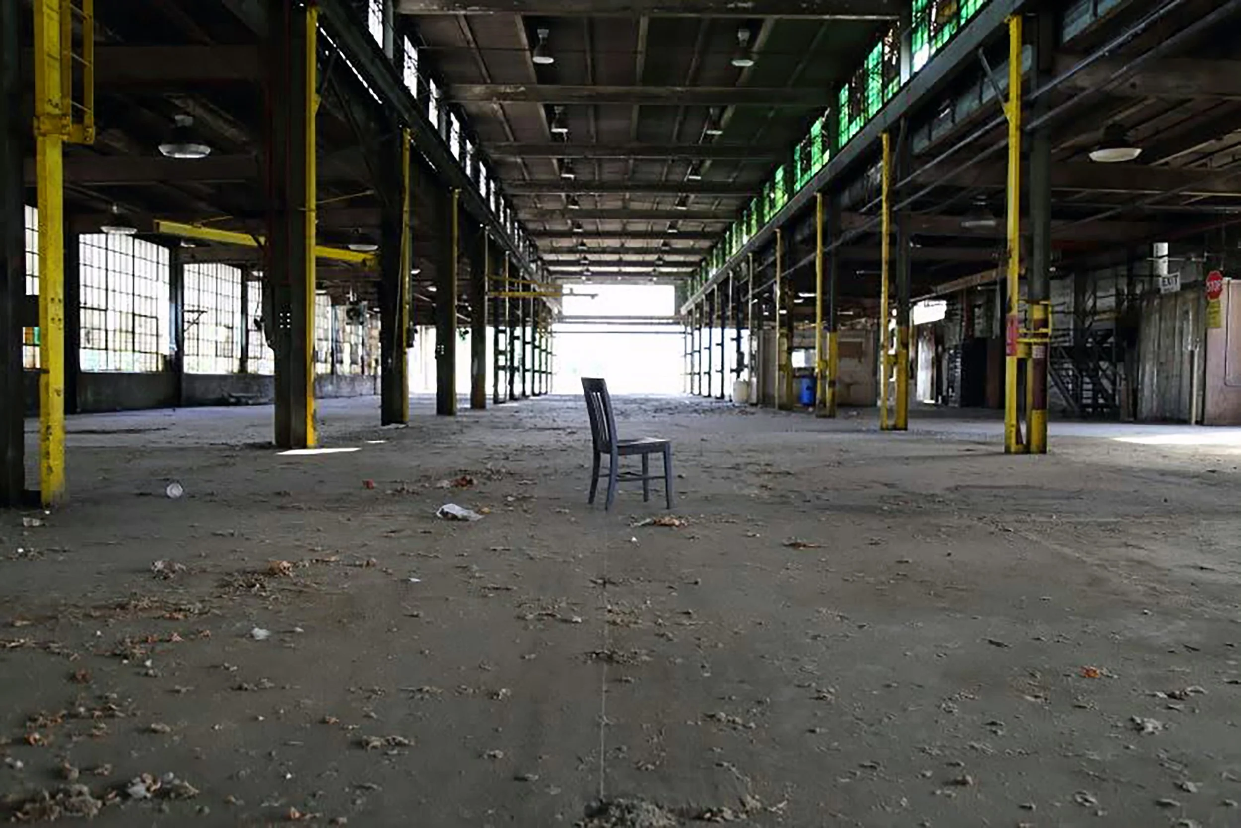 Empty warehouse with scaffolding, yellow support poles, and a single chair on dusty, debris-covered floor.