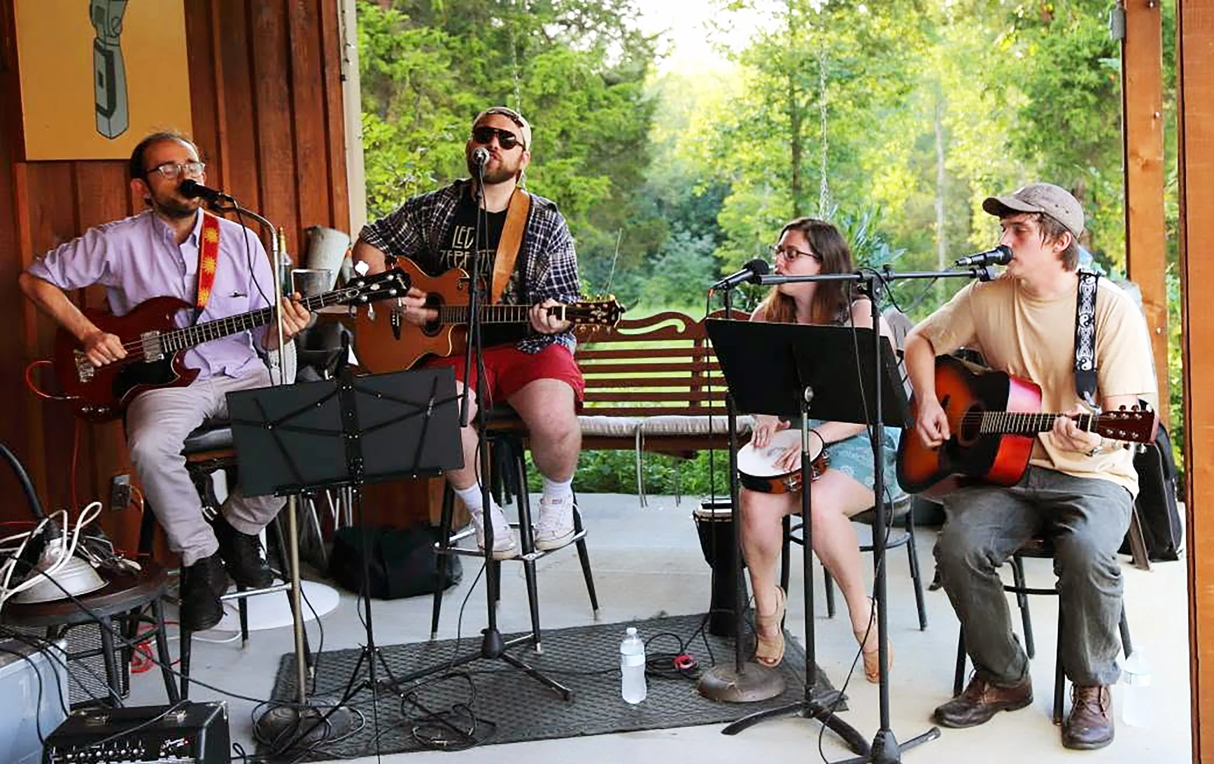 Four musicians performing outdoors on a wooden stage surrounded by green trees, playing guitars and singing into microphones.