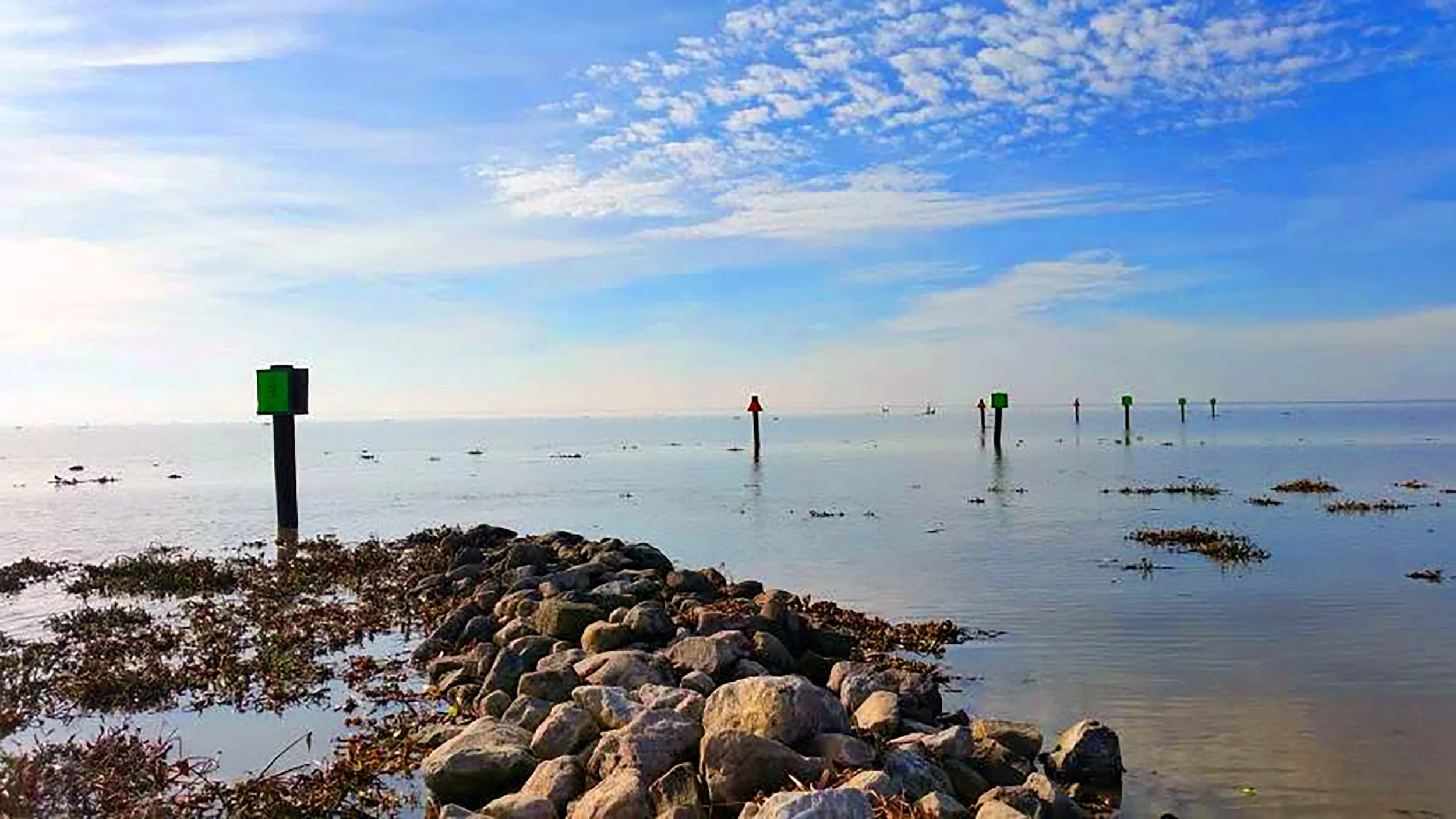 View of a calm sea or lake with a rocky shoreline in the foreground, several green navigation markers in the water, a partly cloudy blue sky, and distant sailboats on the horizon.