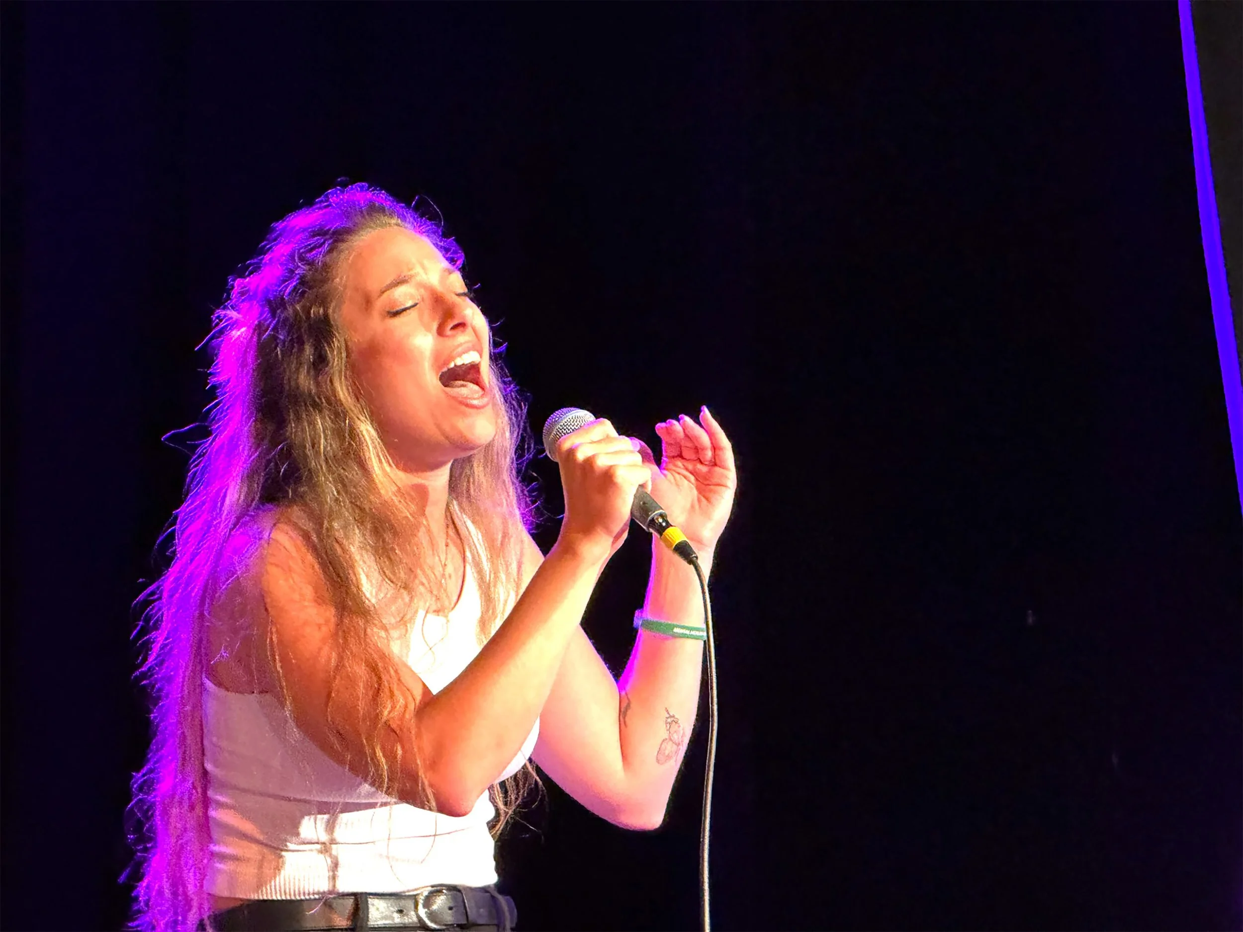 A woman with long, wavy hair singing passionately into a microphone on stage, illuminated by purple stage lights, against a dark background.
