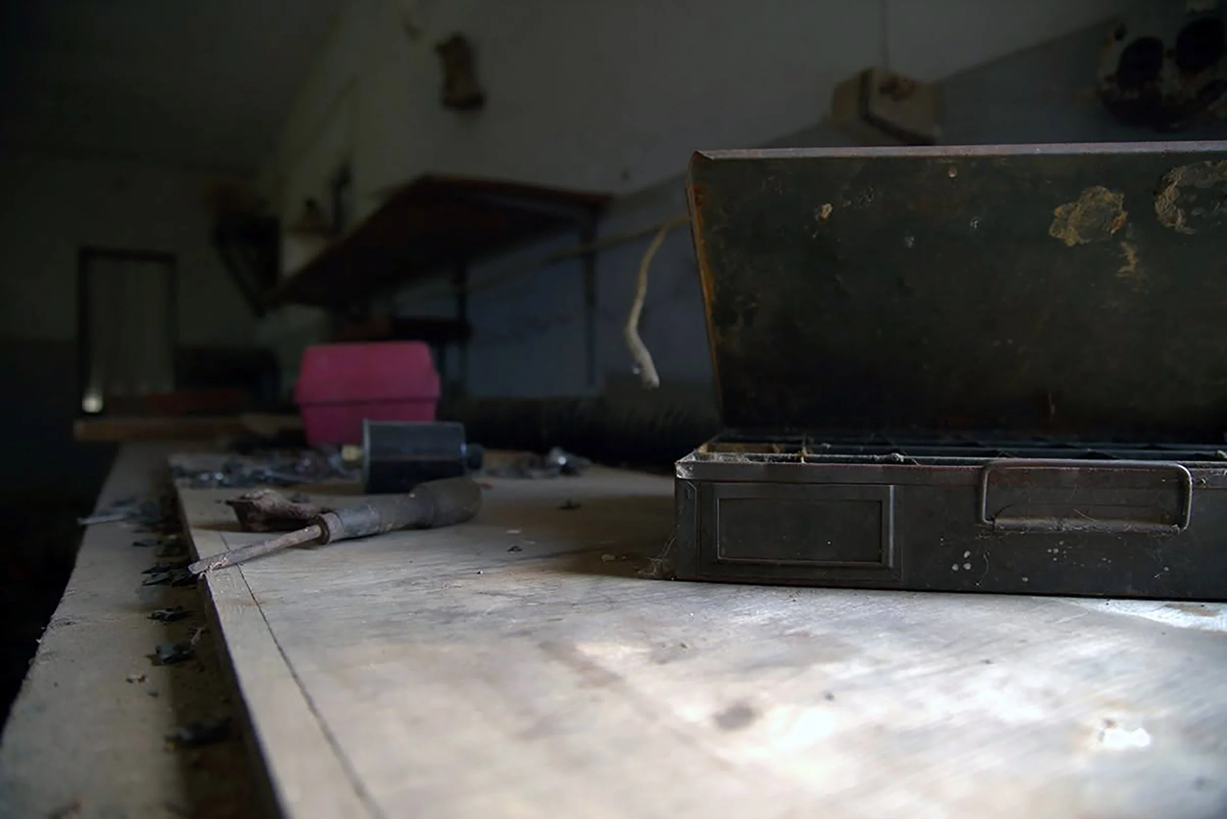 An abandoned, dusty workspace with an open metal box, scattered tools, and debris on a rough wooden surface, with a dark, cluttered background.