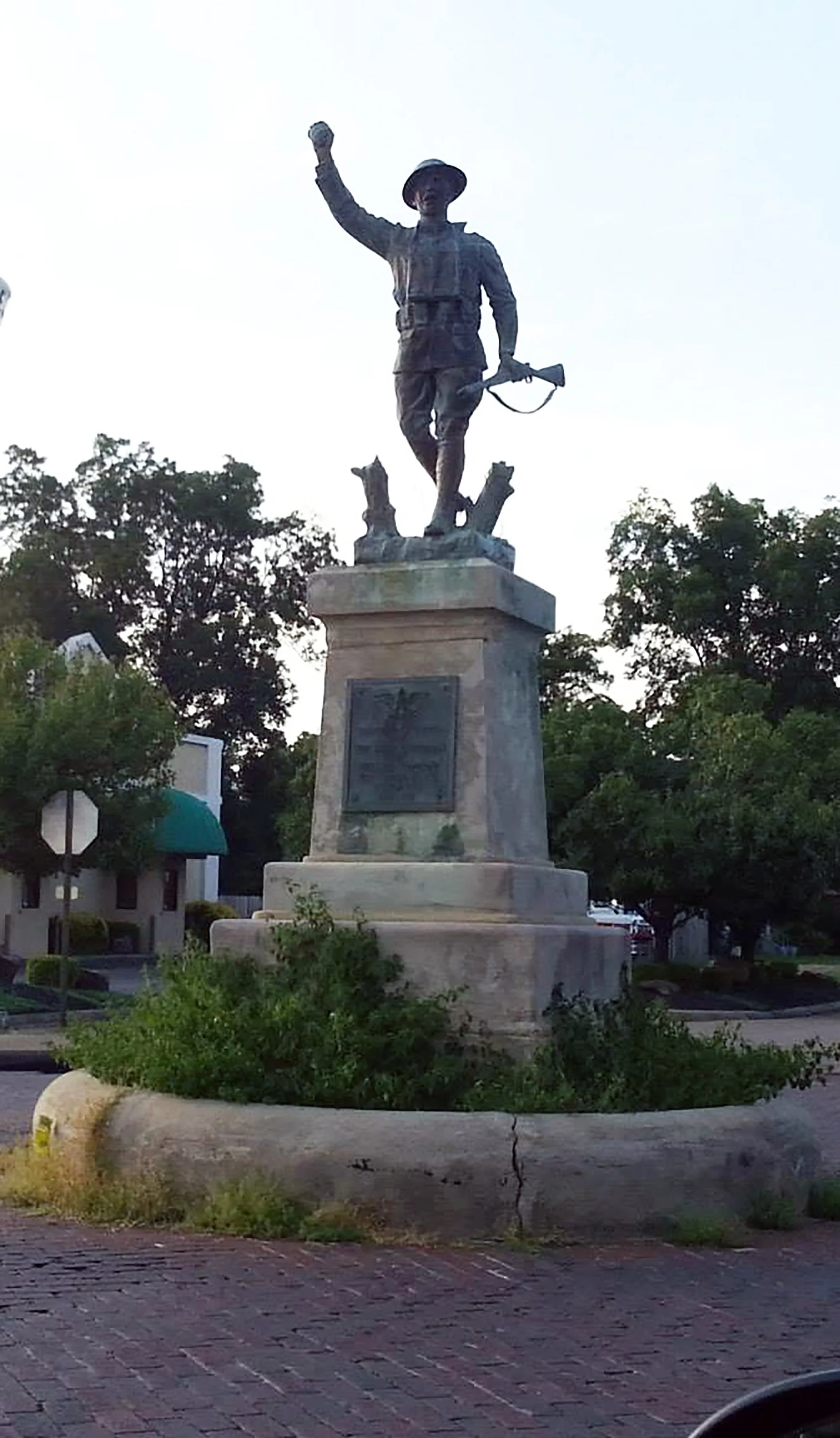 Bronze statue of a soldier holding a rifle, standing on a stone pedestal with a plaque, surrounded by trees and a brick street.