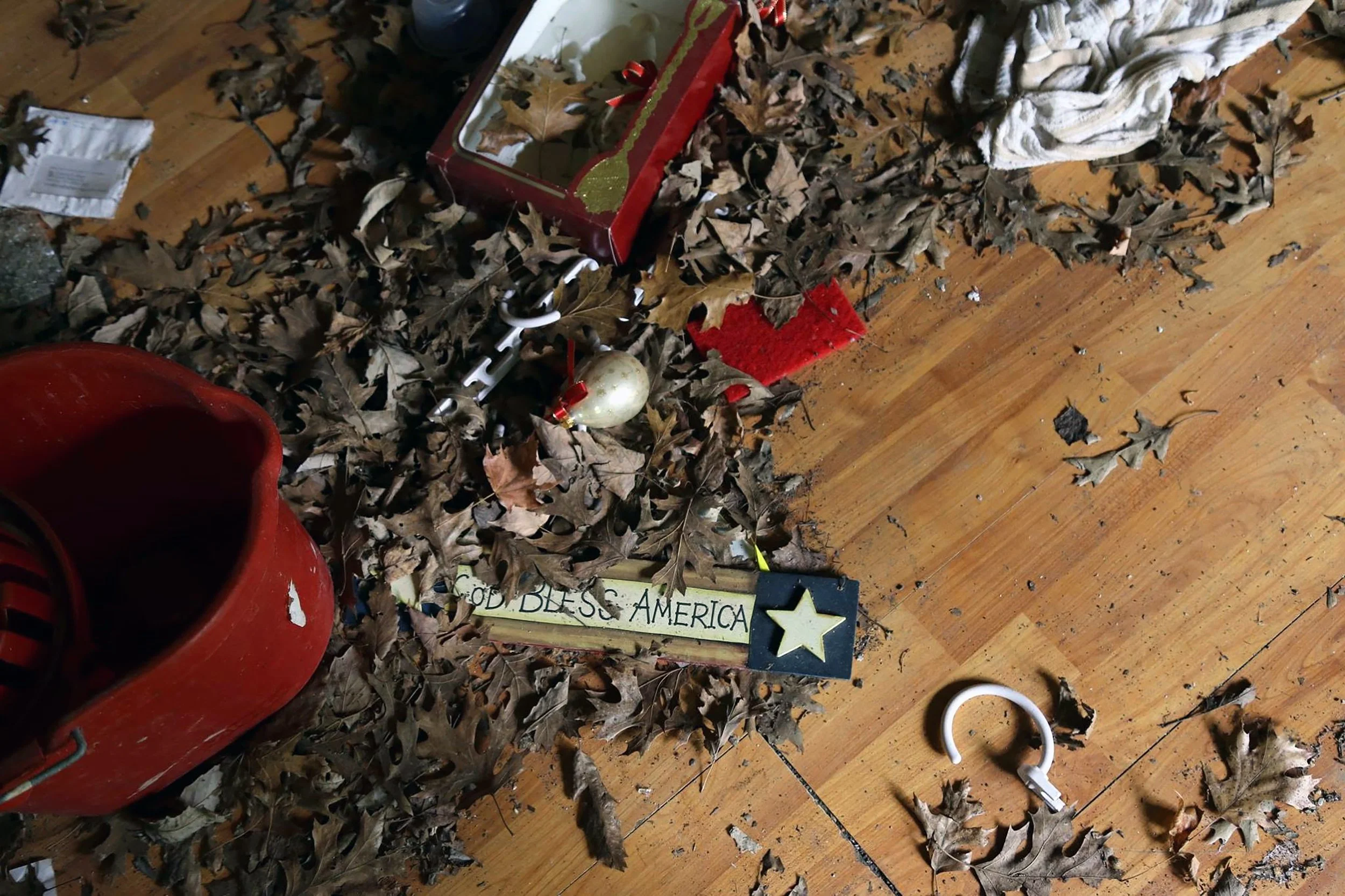 Disorganized clutter on a wooden floor includes a red bucket, a decorative mirror, fallen leaves, a small American flag, a white cloth, a Christmas ornament, and a sign that reads 'God Bless America.'