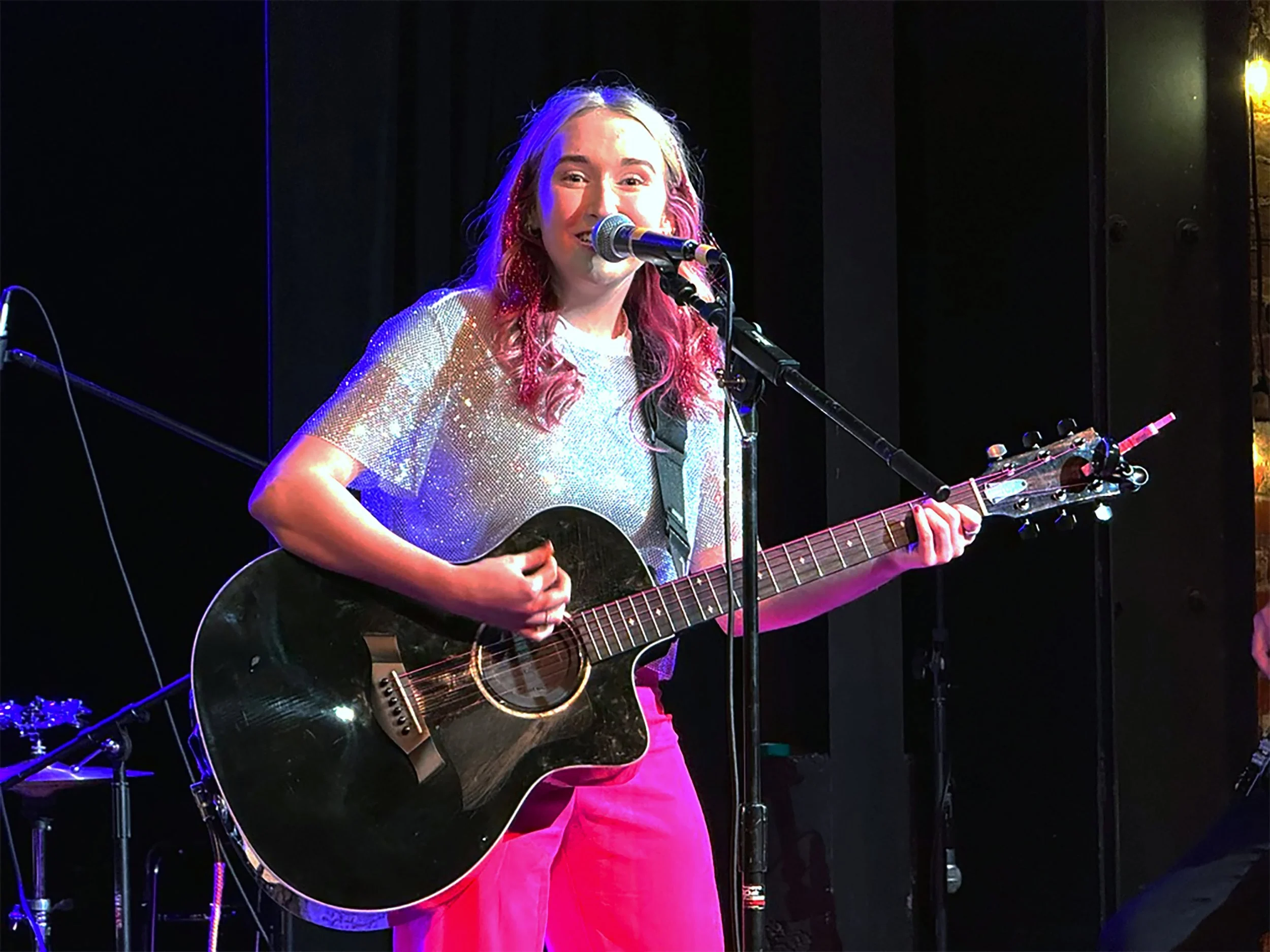 A woman with pink and blonde hair playing an acoustic guitar on stage, smiling and singing into a microphone.