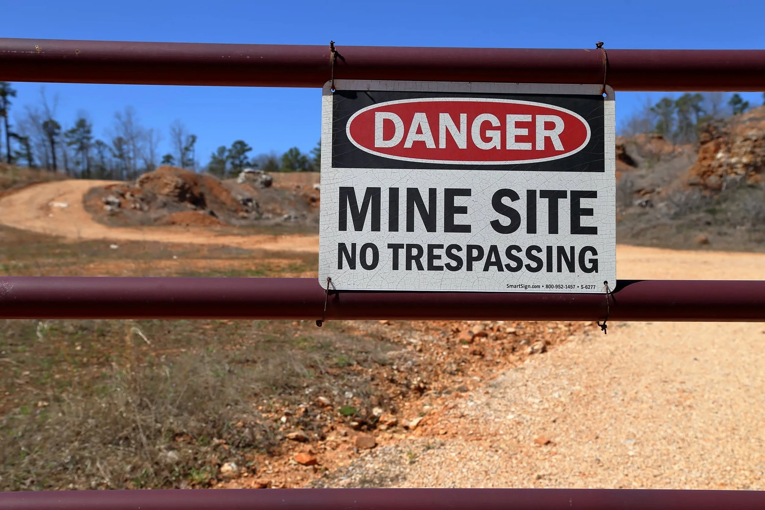 A warning sign hanging on a metal gate that reads 'DANGER MINE SITE NO TRESPASSING' with a dirt path and barren landscape in the background.