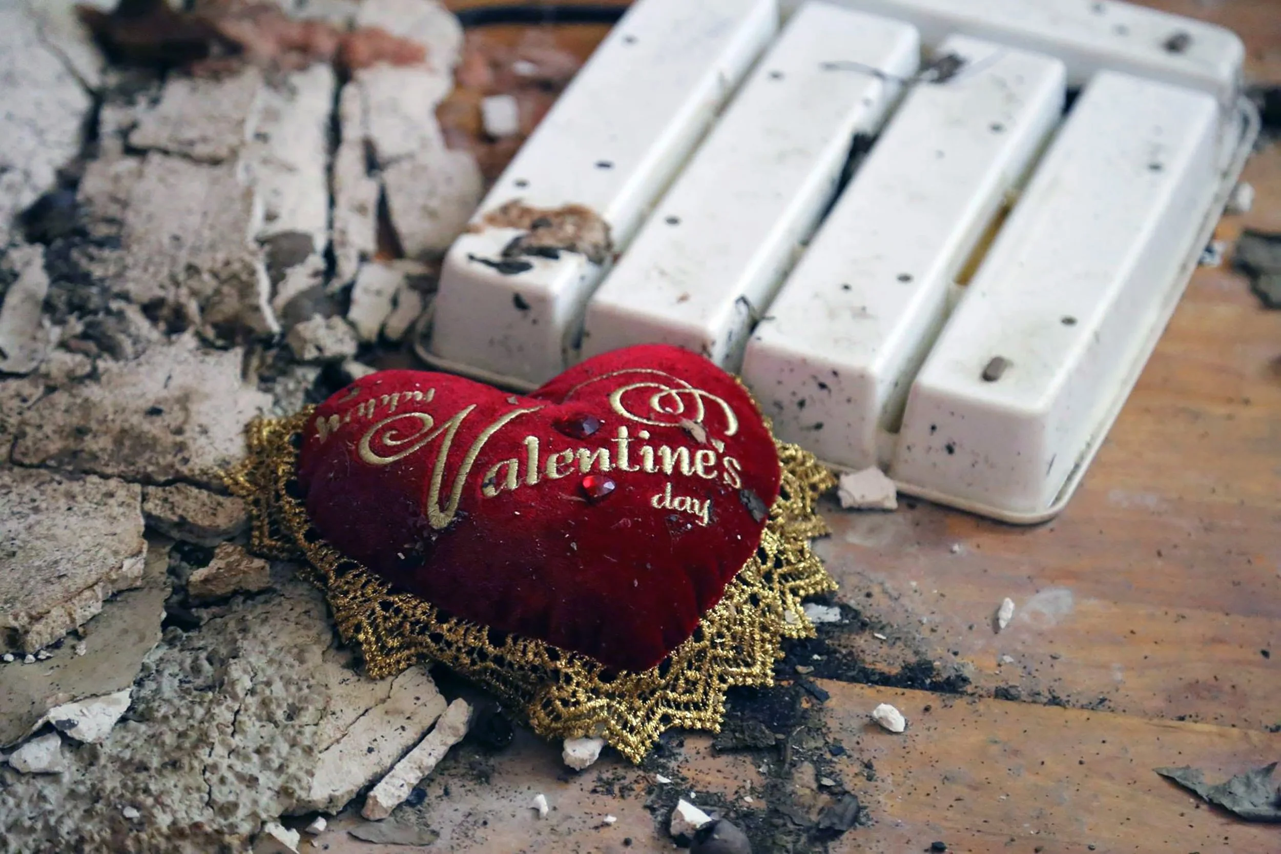 A velvety red Valentine's Day heart-shaped ornament, embroidered with gold thread, surrounded by gold tinsel, lying on dirty, ashen ground near broken white ceramic pieces and ash.