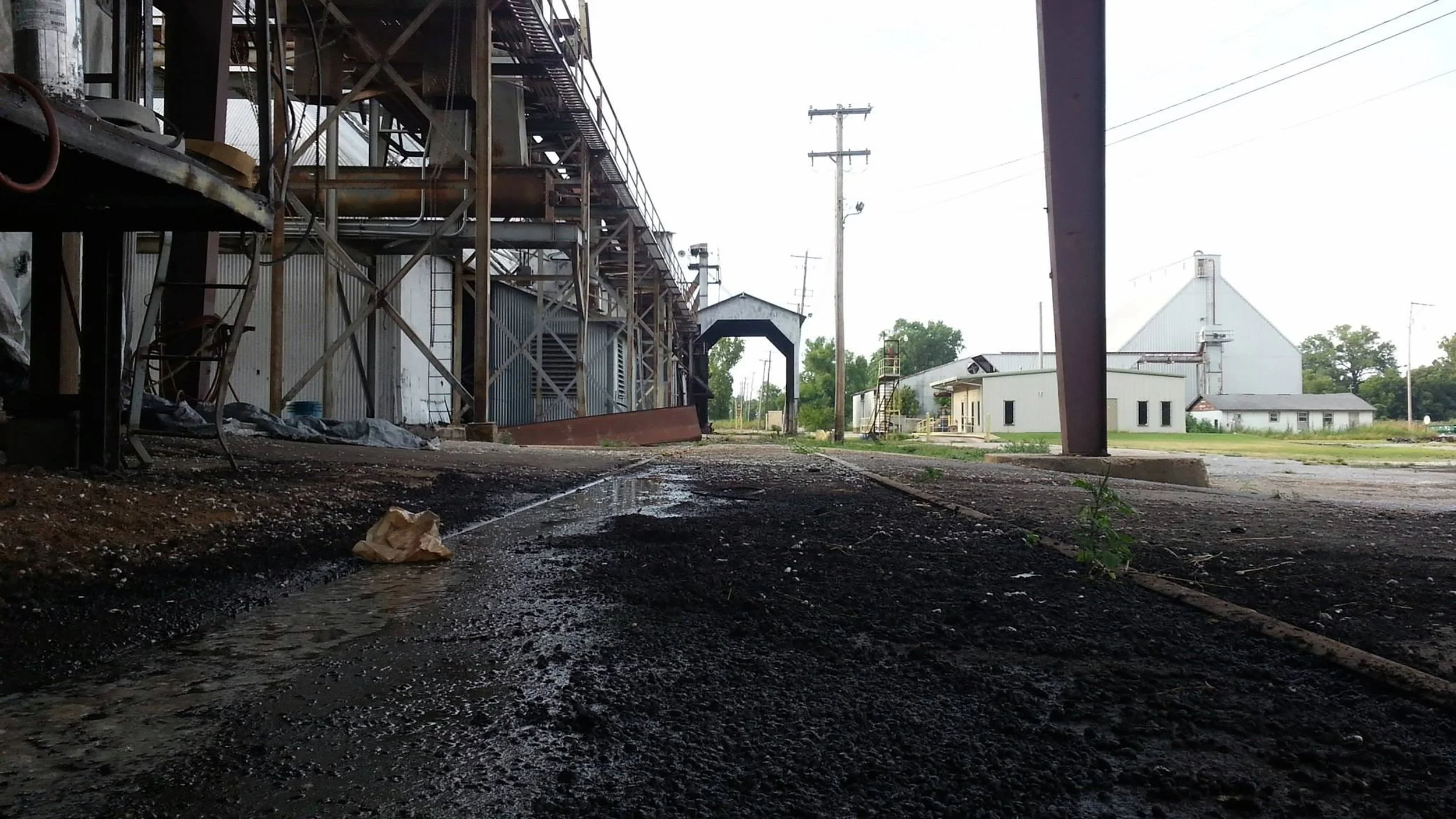 View of an industrial area with metal buildings, power lines, and a muddy ground with water puddles.