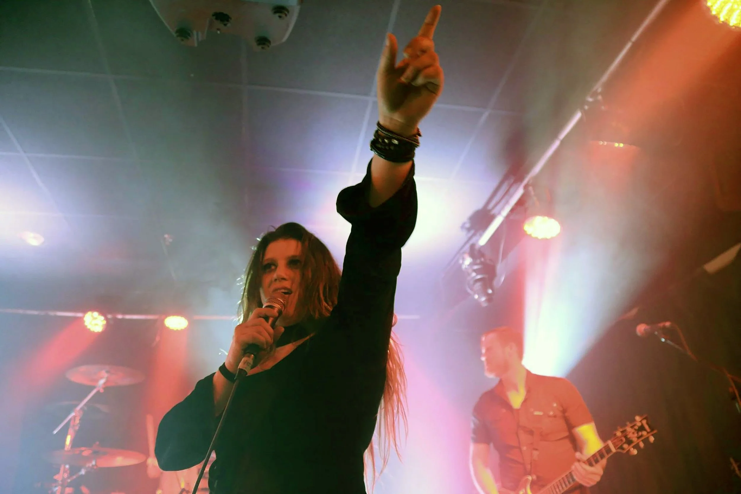 A female singer is performing on stage with a microphone in her right hand and her left arm raised. She has long hair, and is surrounded by colorful stage lights with a band in the background playing instruments.