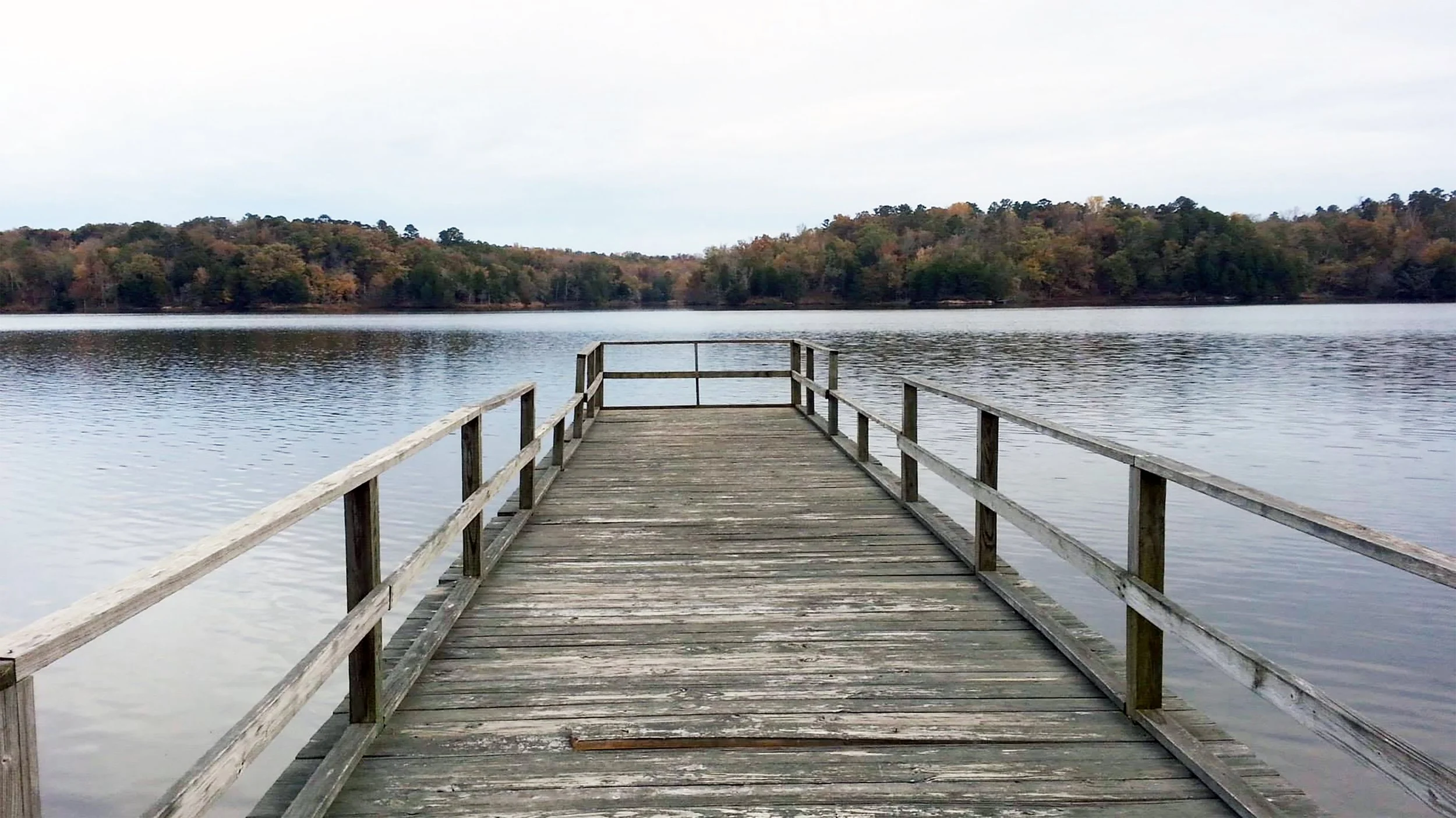A wooden dock extending into a calm lake surrounded by trees with autumn foliage.