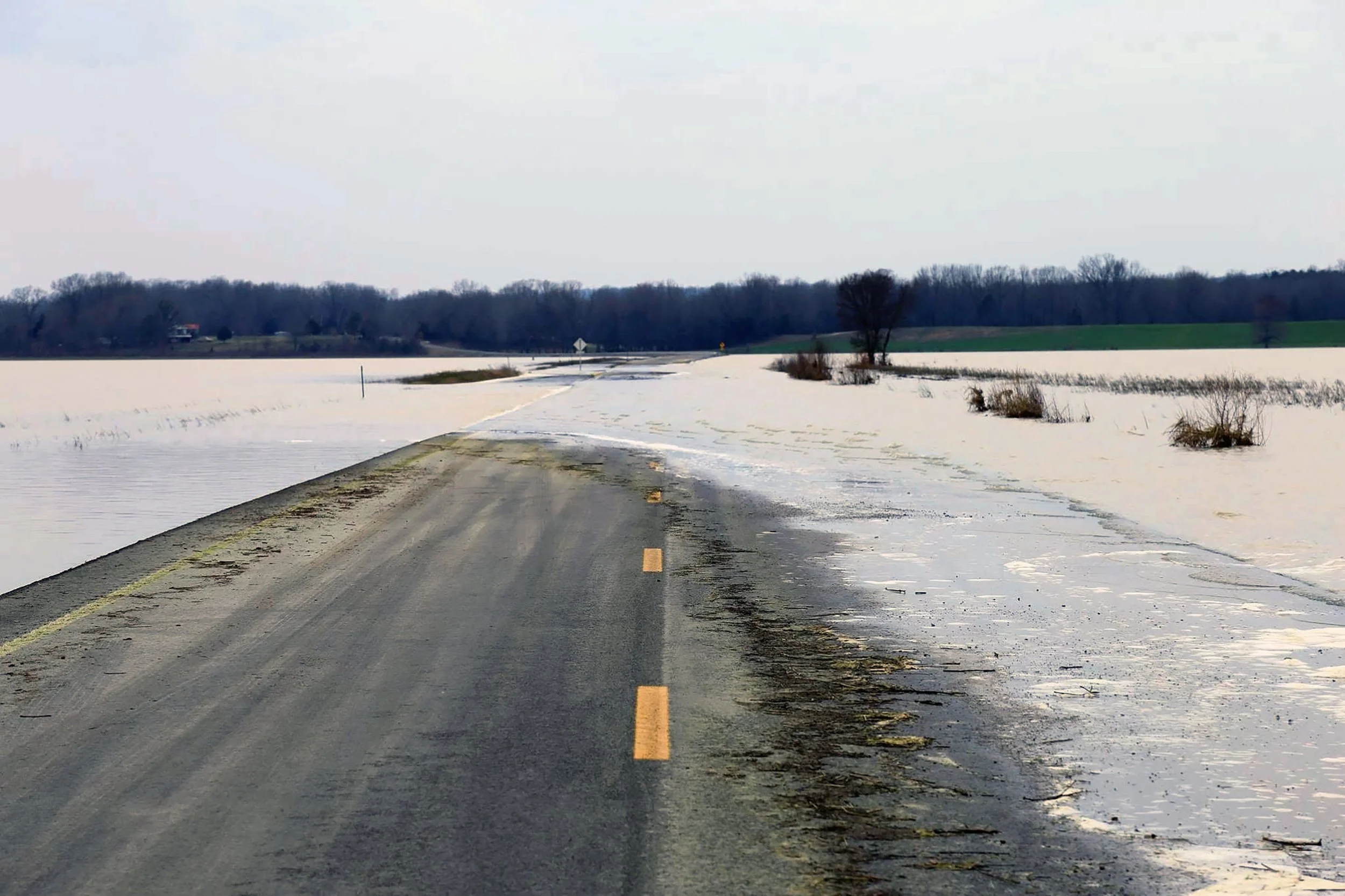 A flooded road with partially submerged asphalt, surrounded by snow and wet grass, with trees and fields in the background under an overcast sky.