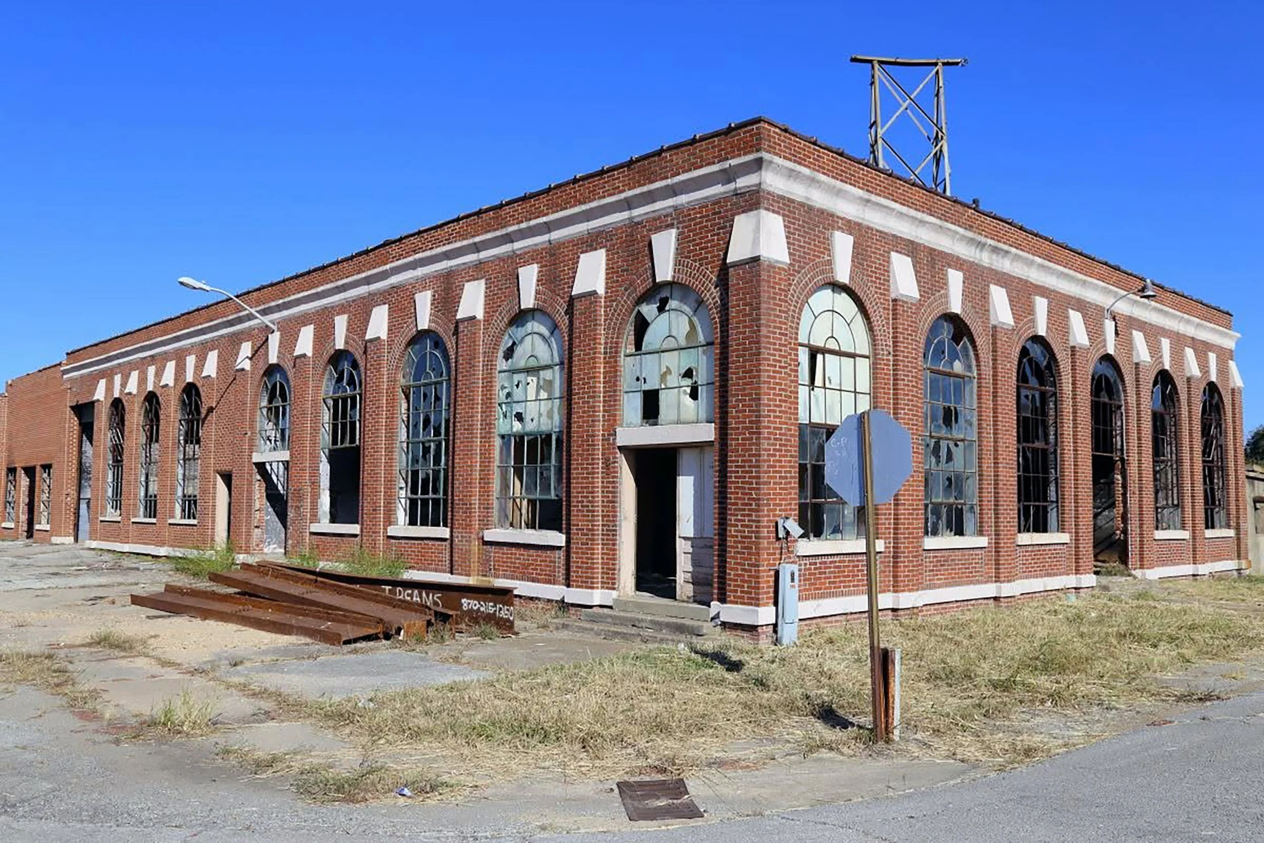 An old brick building with broken windows and boarded-up entrances, situated on a mostly empty lot with some patches of grass and scattered debris, under a clear blue sky.