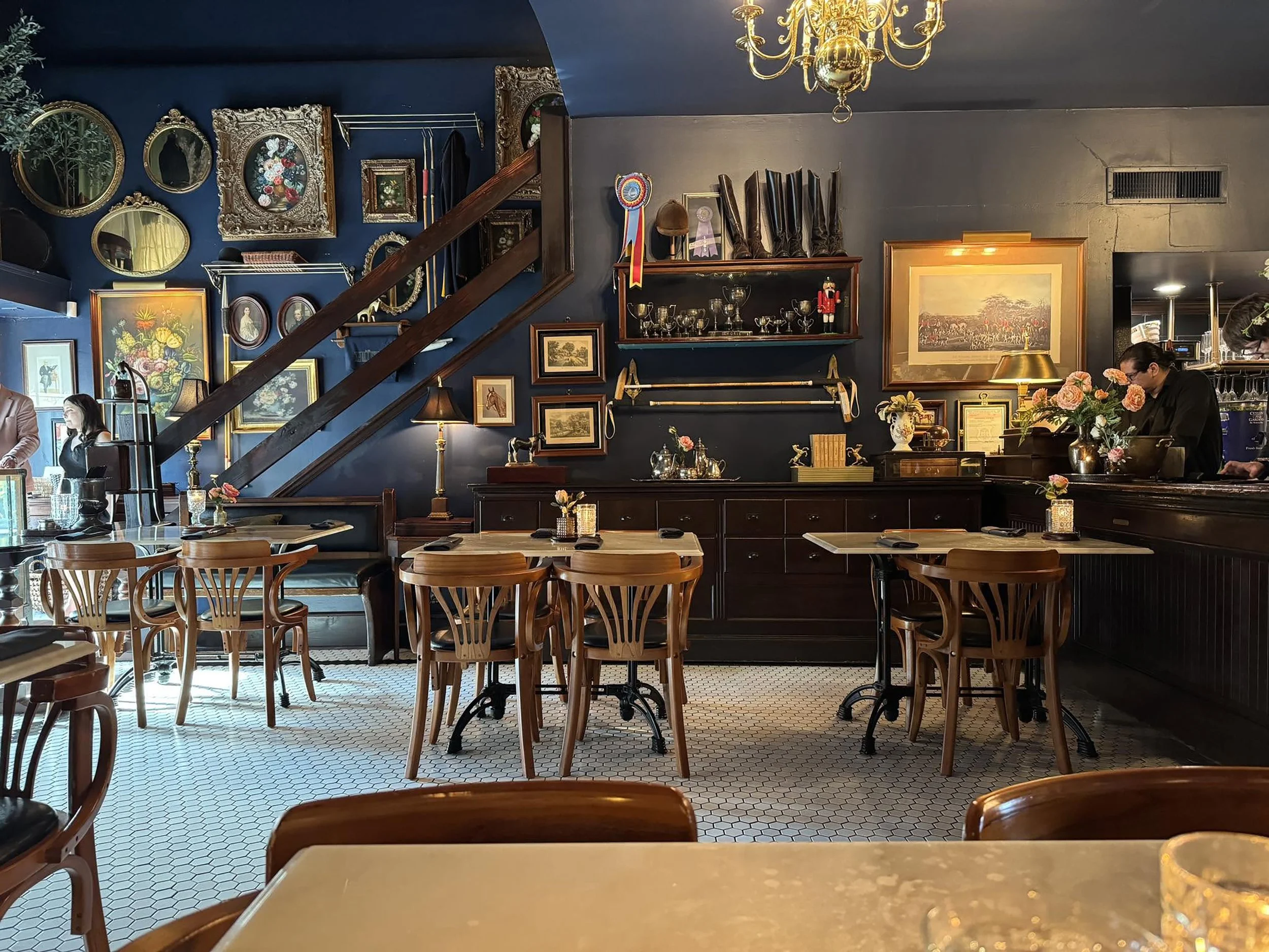 Interior of a vintage-style restaurant with blue and gray walls decorated with framed artwork, mirrors, and trophies. Wooden tables and chairs are arranged on a white hexagonal tile floor. A person wearing a black jacket and glasses is in the backgro