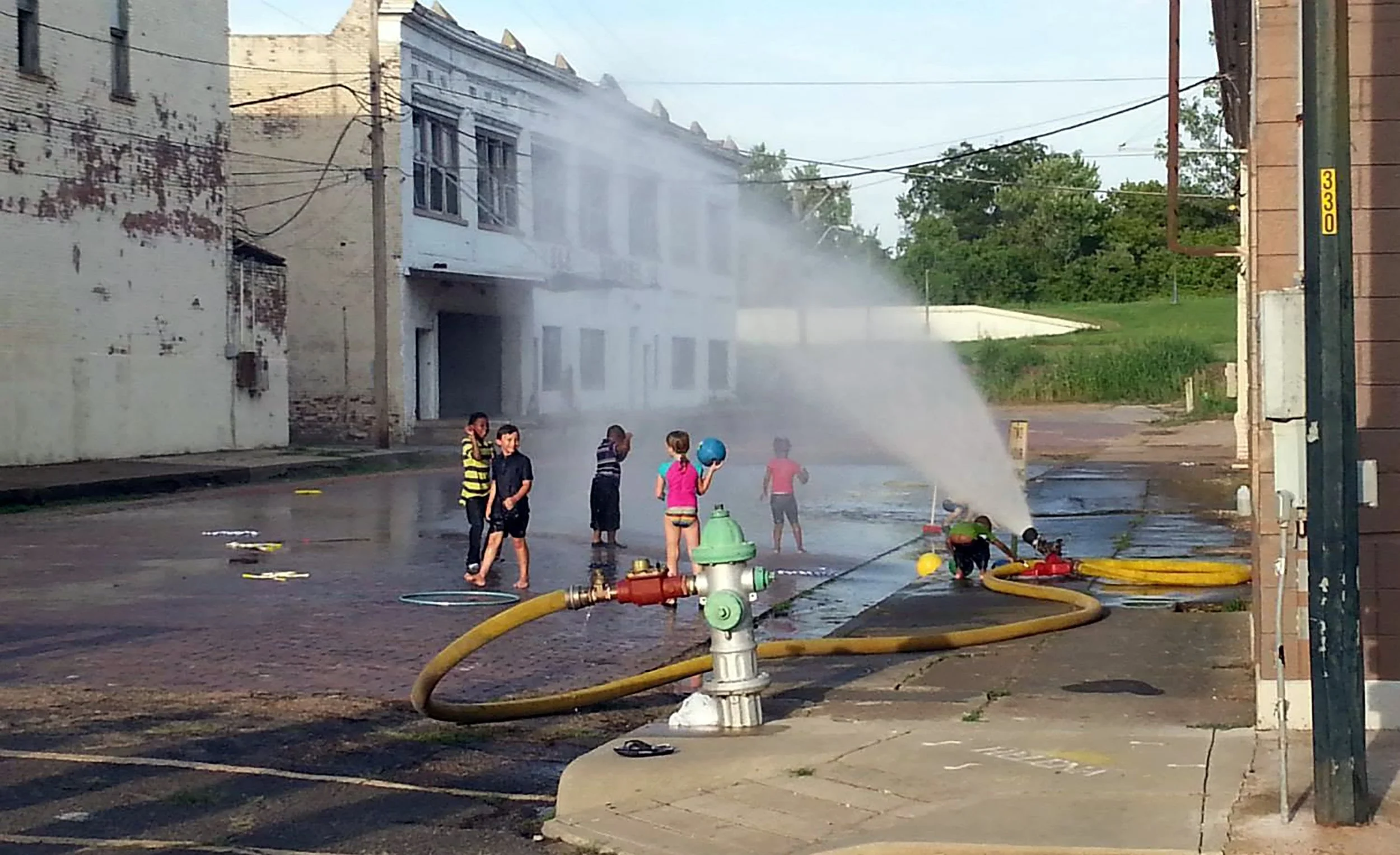 Children playing in a street with water spraying from fire hoses and sprinklers, on a sunny day.