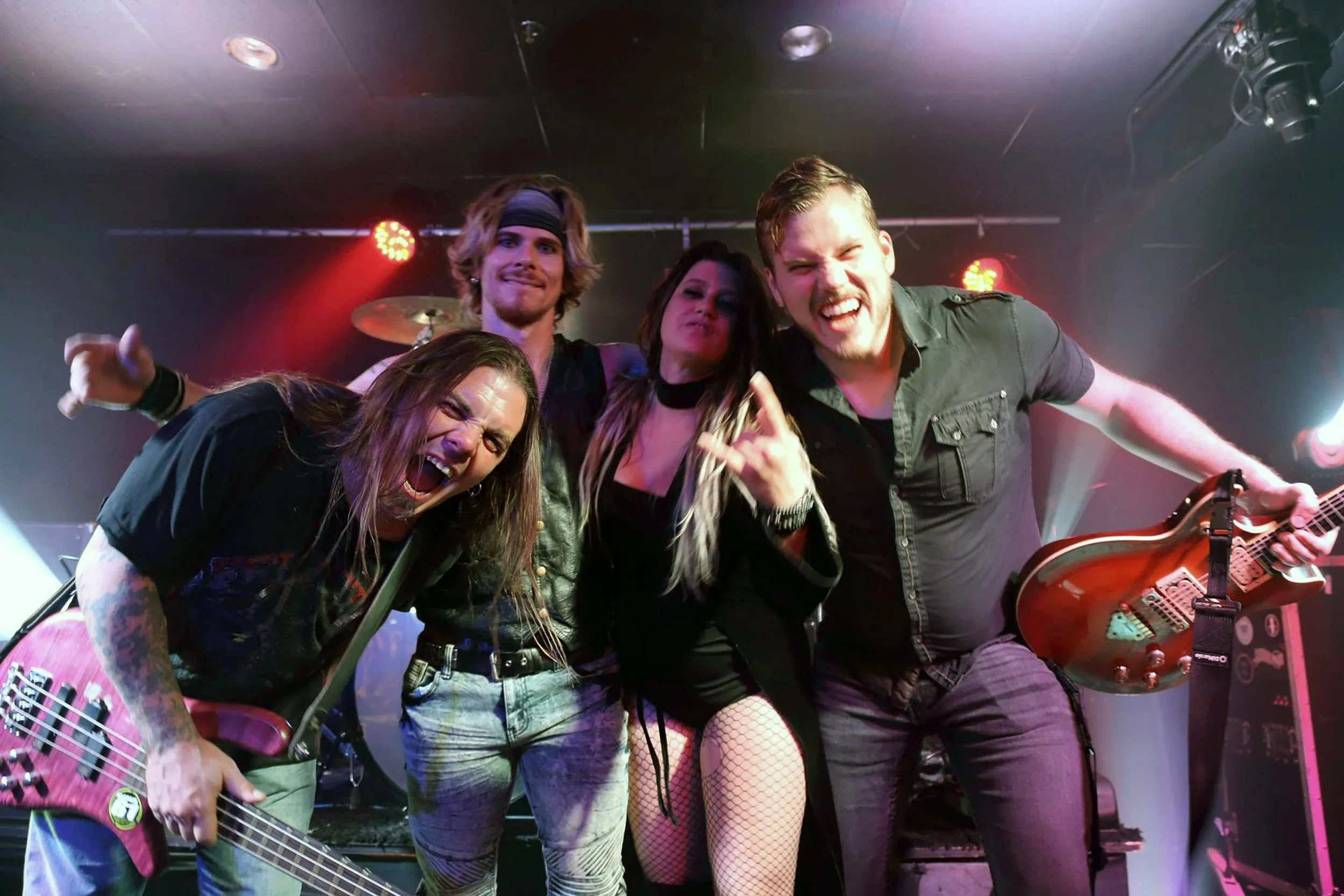 A group of five musicians performing on stage with colorful lighting. One woman is singing with a tattooed arm holding a pink electric guitar, and a man is playing an acoustic guitar while smiling. The background has a drum set and stage lights.