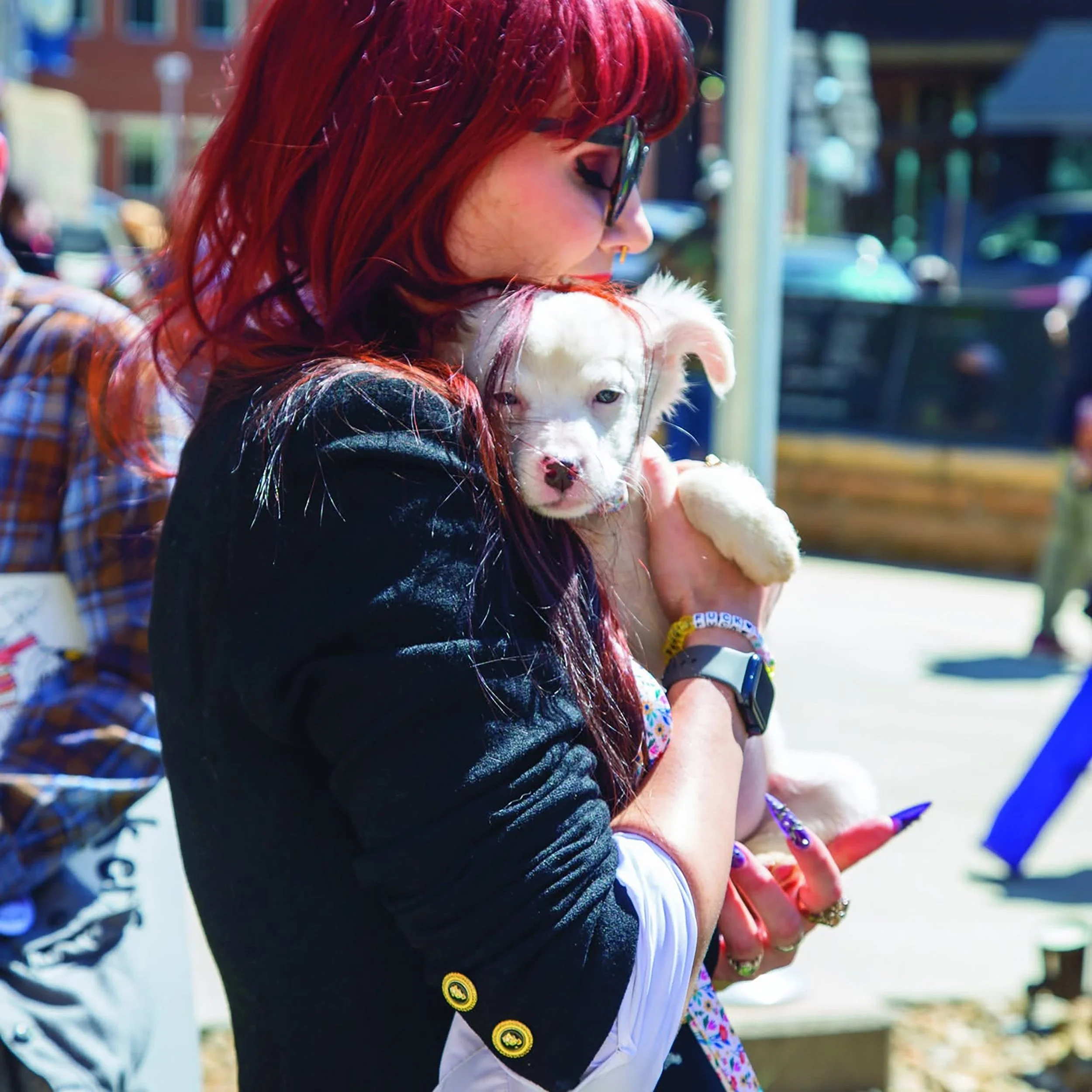 A woman with long red hair, wearing glasses, a black jacket, and colorful jewelry, is holding a white puppy with blue eyes close to her face outdoors on a sunny day.