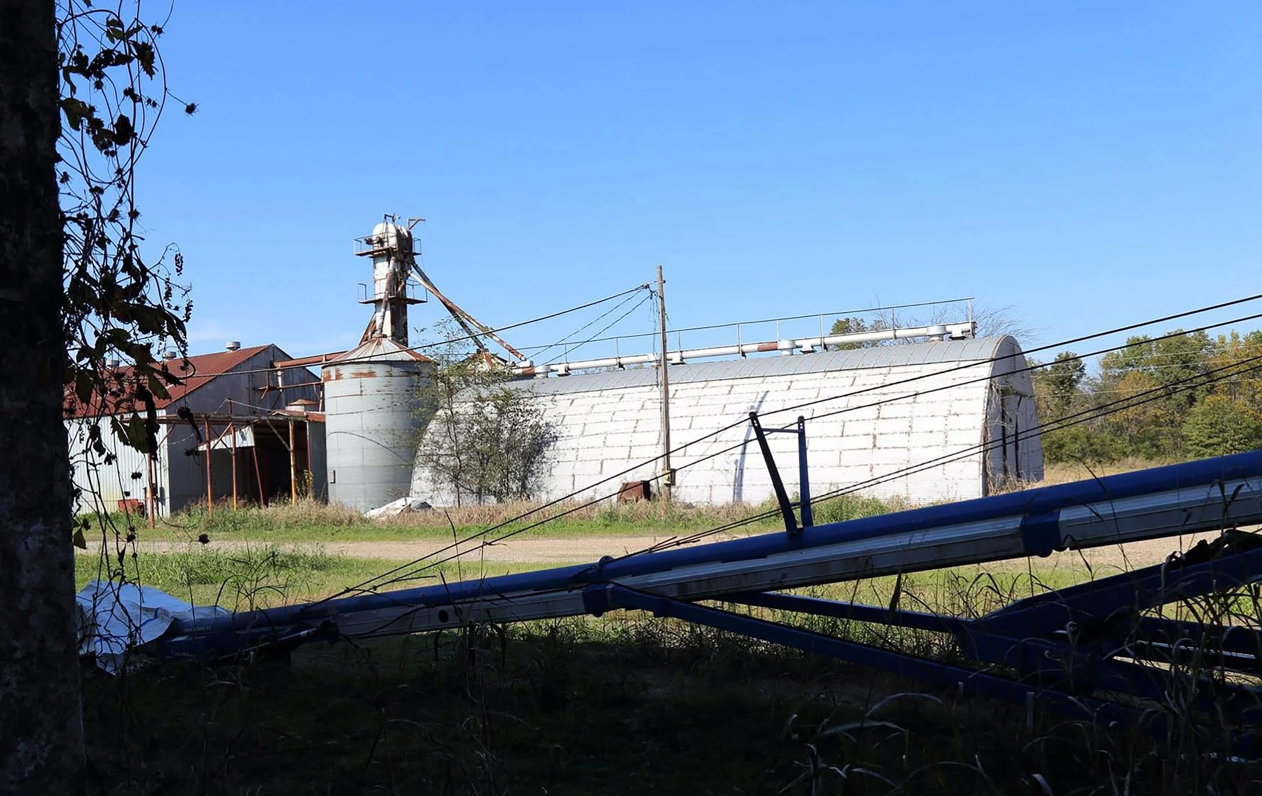 An old, weathered barn and silo with rusted metal and a curved roof, surrounded by power lines and trees, under a clear blue sky.