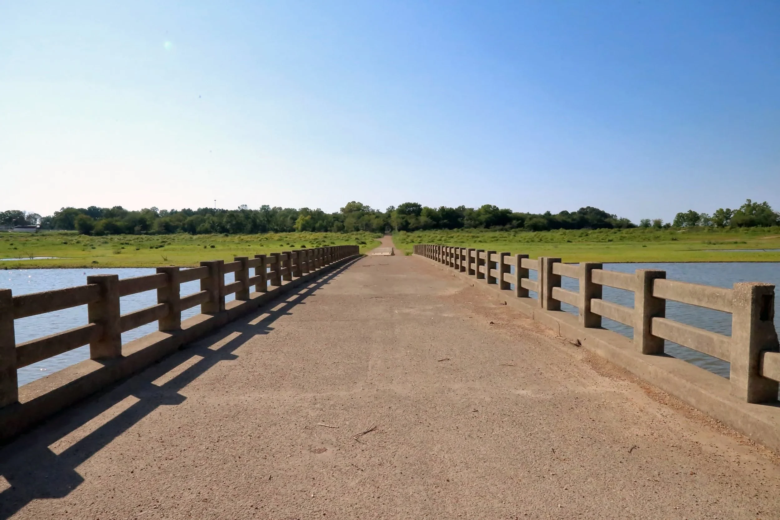 A concrete bridge extends over a body of water, with a gravel path leading into a green, tree-filled landscape under a clear blue sky.