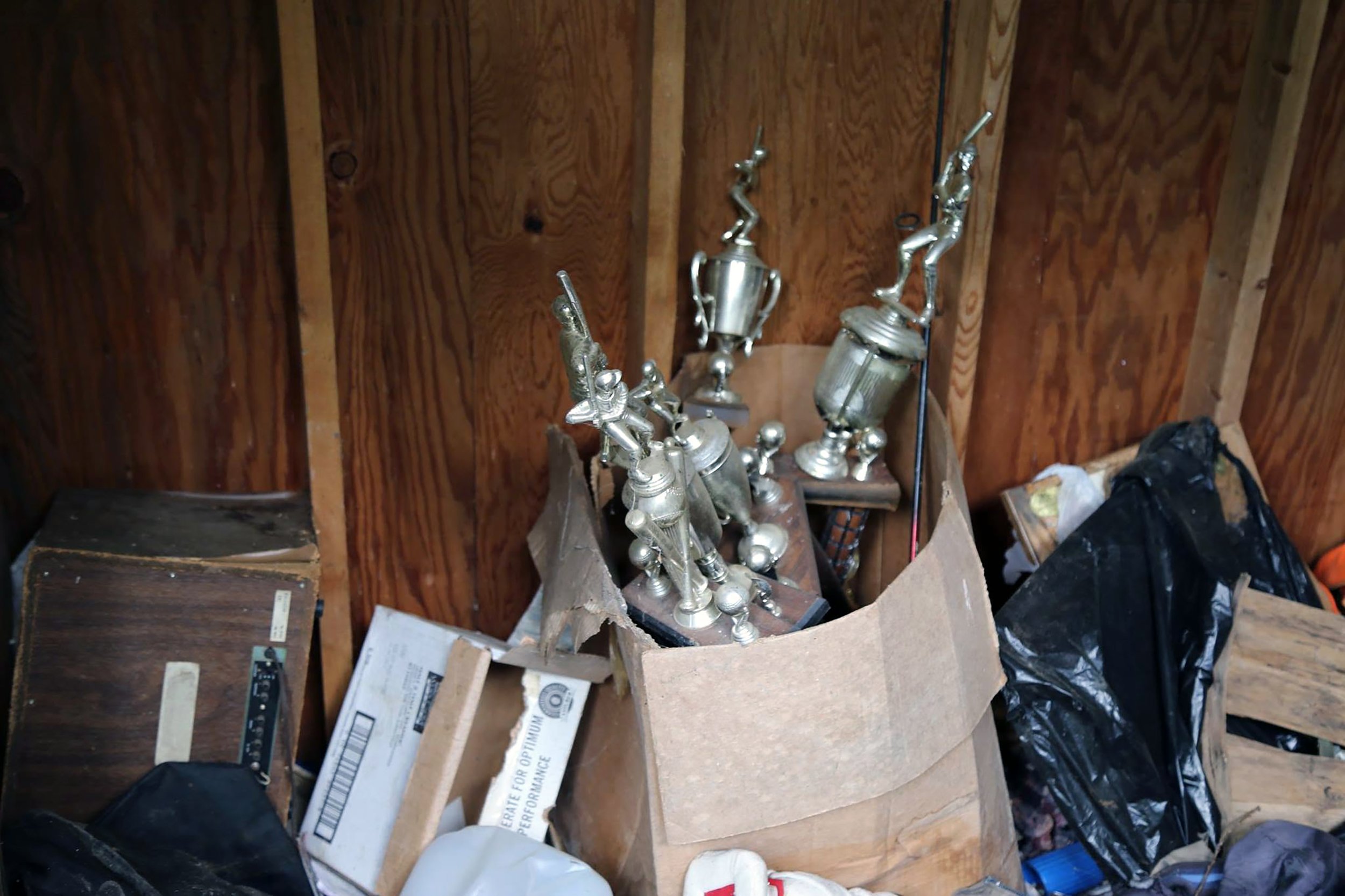 A cluttered storage space with wooden walls and a collection of trophies placed in a cardboard box surrounded by scattered papers, plastic bags, and various items.