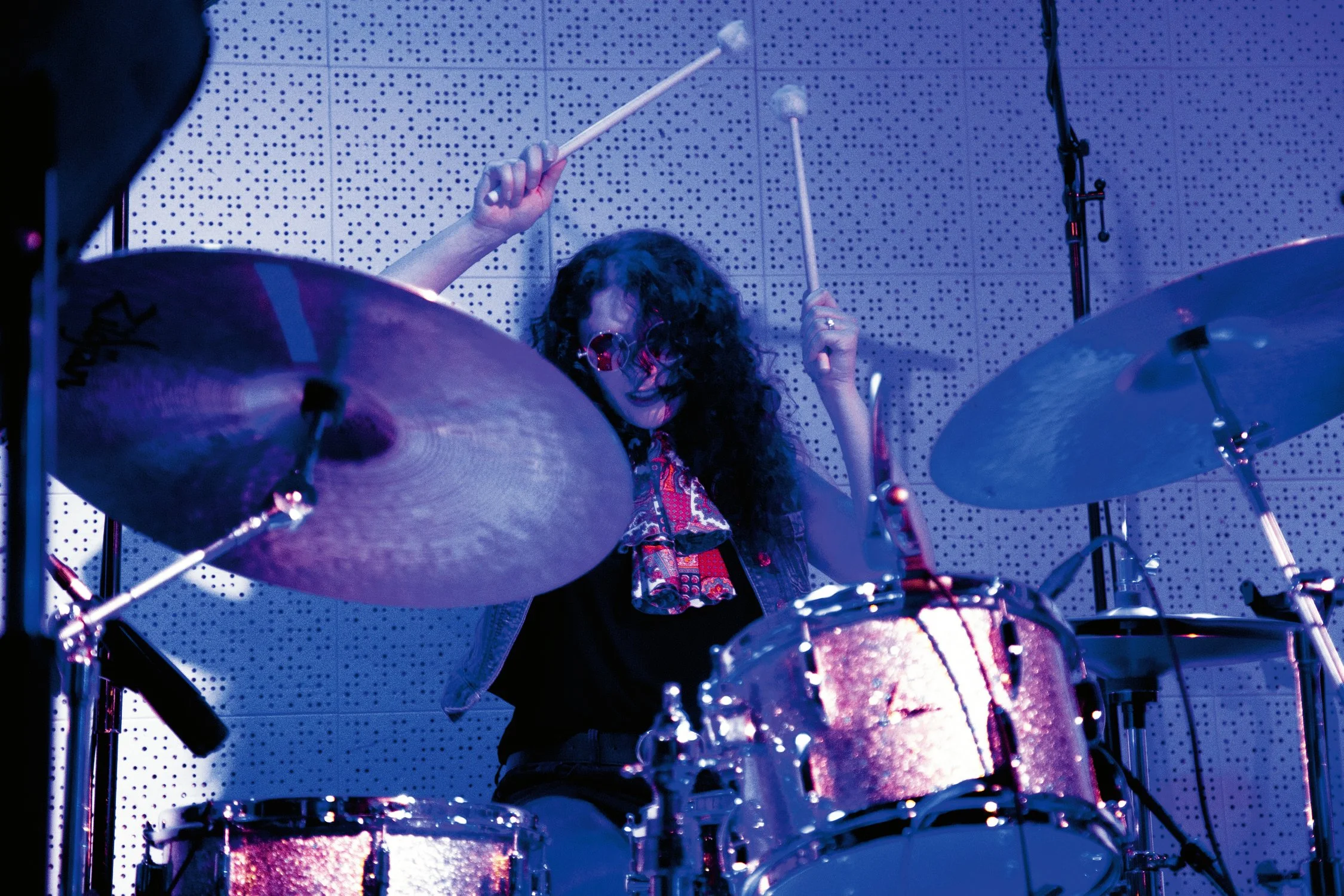 A woman with curly hair playing a drum set in a music studio, holding drumsticks with both hands, surrounded by drums and cymbals.