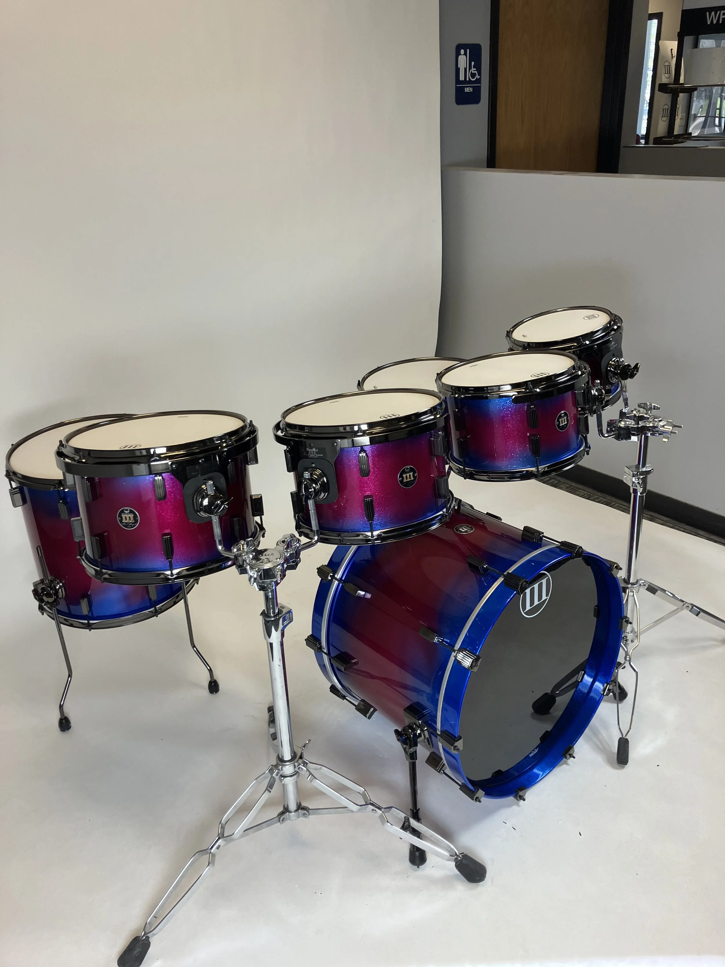 A drum set with pink and blue gradient finish on a white floor, with a white wall in the background and a partially visible doorway.